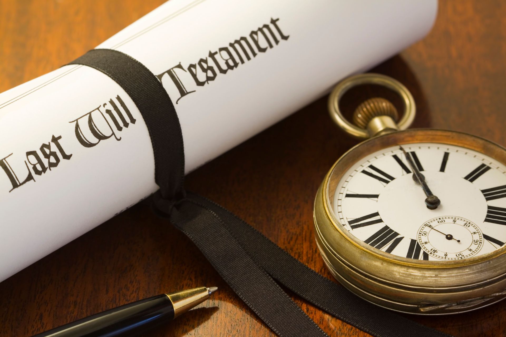 Rolled Last Will and Testament scroll with black ribbon, pen, and pocket watch on a wooden surface.