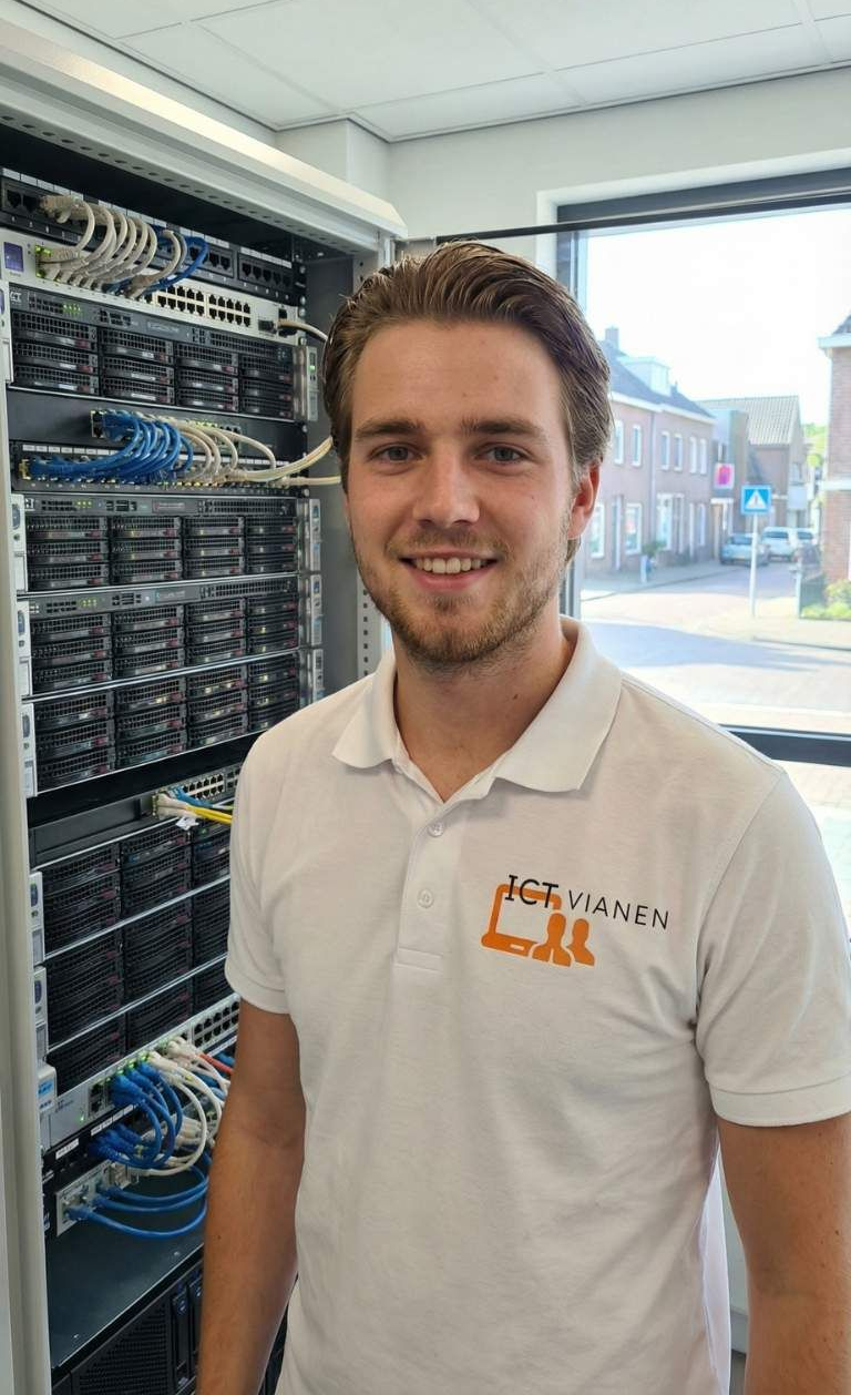 Een man in een wit poloshirt glimlacht voor een serverkast. Daglicht stroomt door een raam naar binnen.