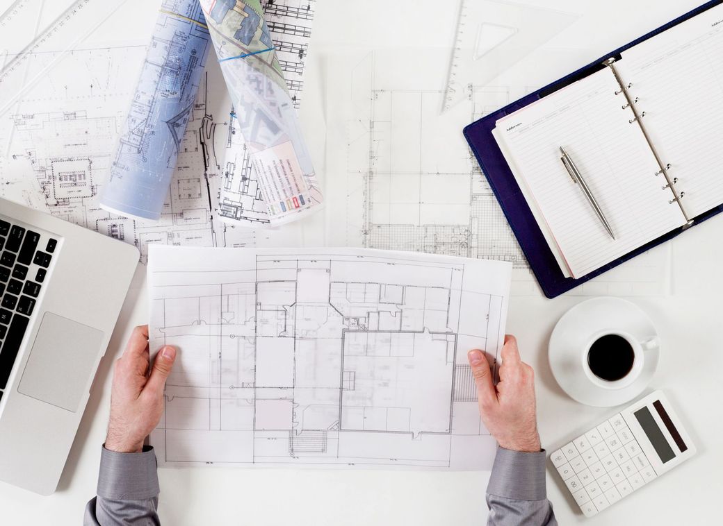 Architetto che esamina le planimetrie a una scrivania con computer portatile, caffè, calcolatrice e taccuino.