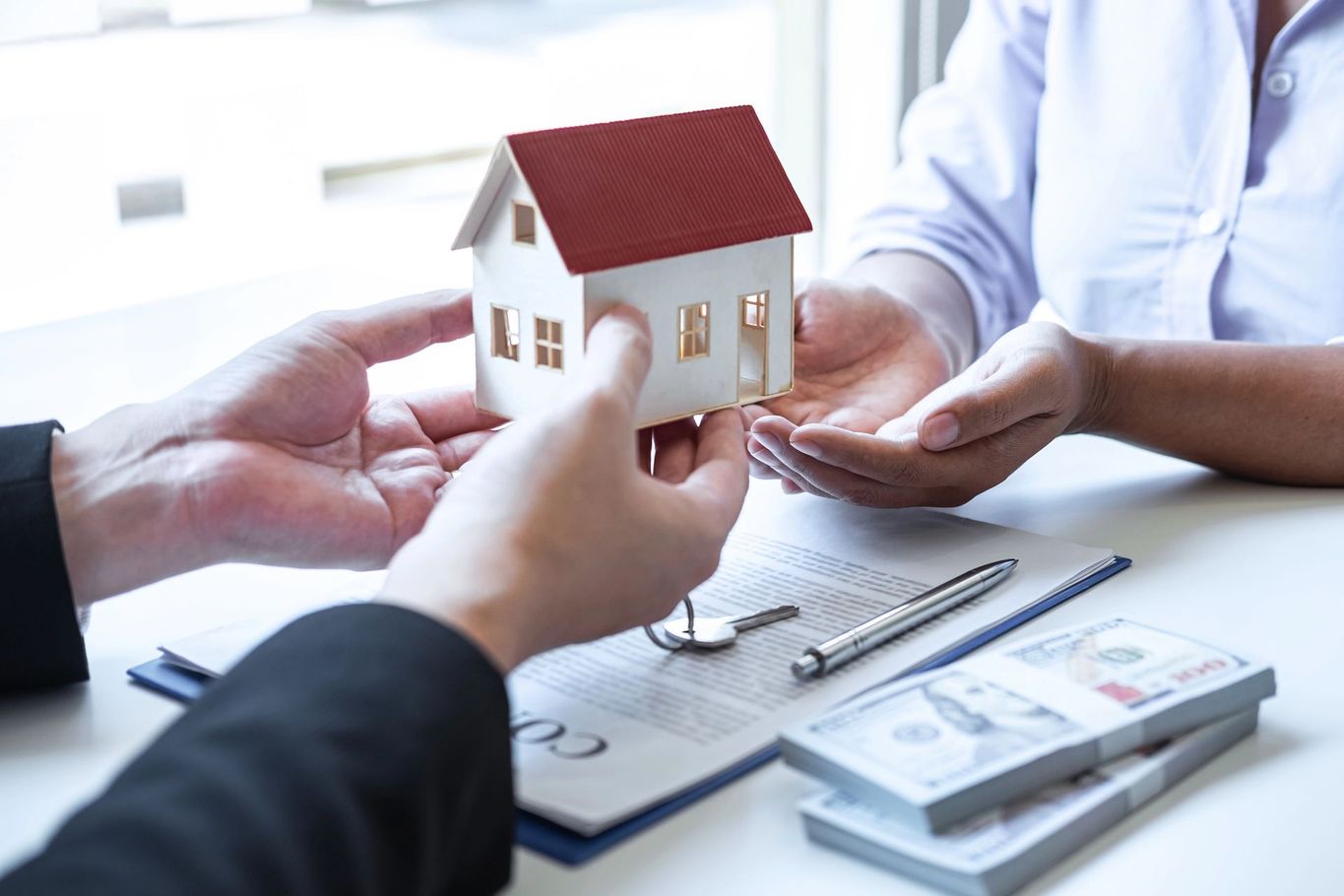 Mani che si scambiano un modellino di casa su una scrivania con documenti finanziari e contanti