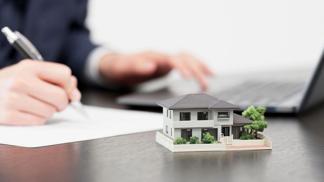 Mani che scrivono su un foglio accanto a un modellino di casa e un computer portatile su una scrivania.