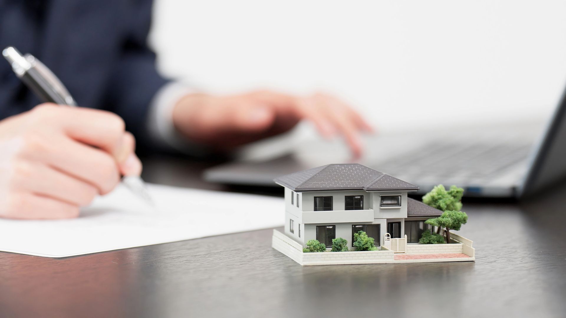 Scrittura a mano su un foglio accanto a un modellino di casa in miniatura e un computer portatile su una scrivania