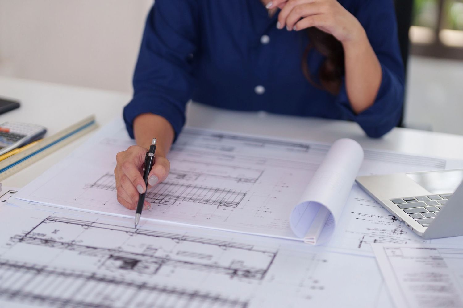 Una persona sta disegnando progetti architettonici su una scrivania, con planimetrie arrotolate e fogli di carta nelle vicinanze.