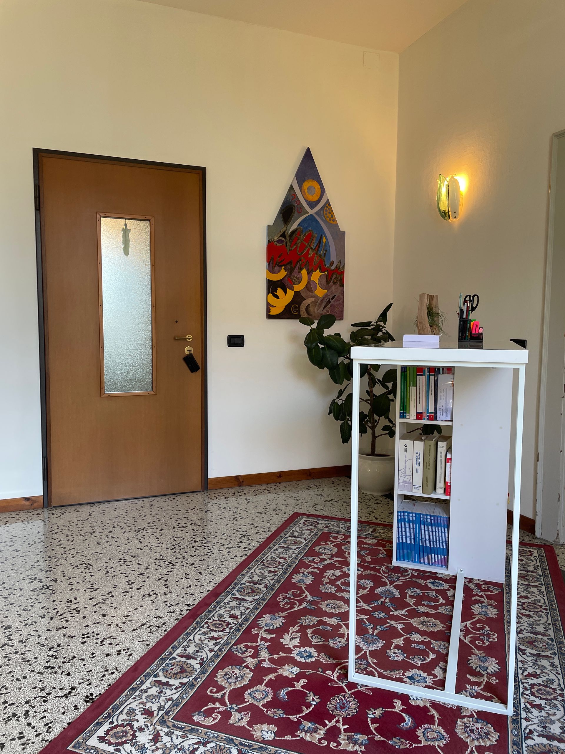 Corridoio tranquillo con porta in legno, tappeto a motivi geometrici, mensola bianca, lampada da parete e quadro decorativo.