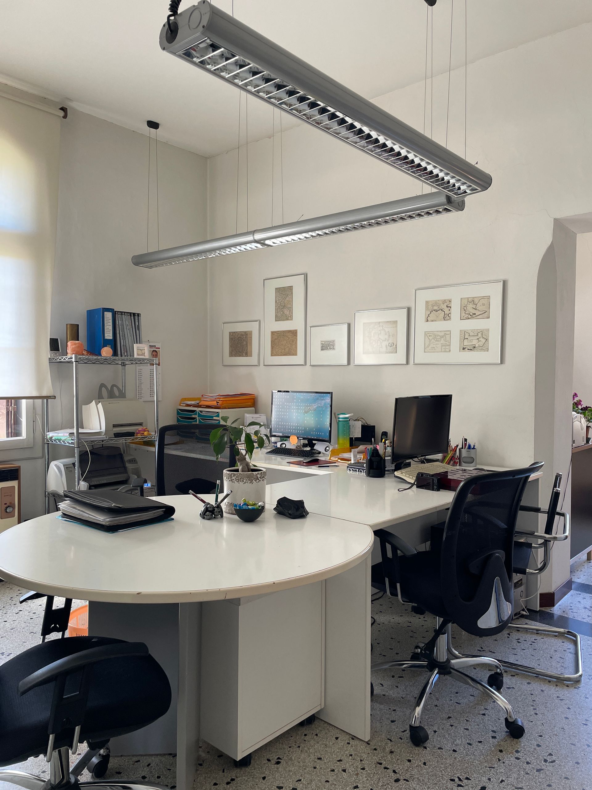 Ufficio moderno con scrivanie, computer, sedie e opere d'arte alle pareti, illuminato da lunghe lampade a sospensione.