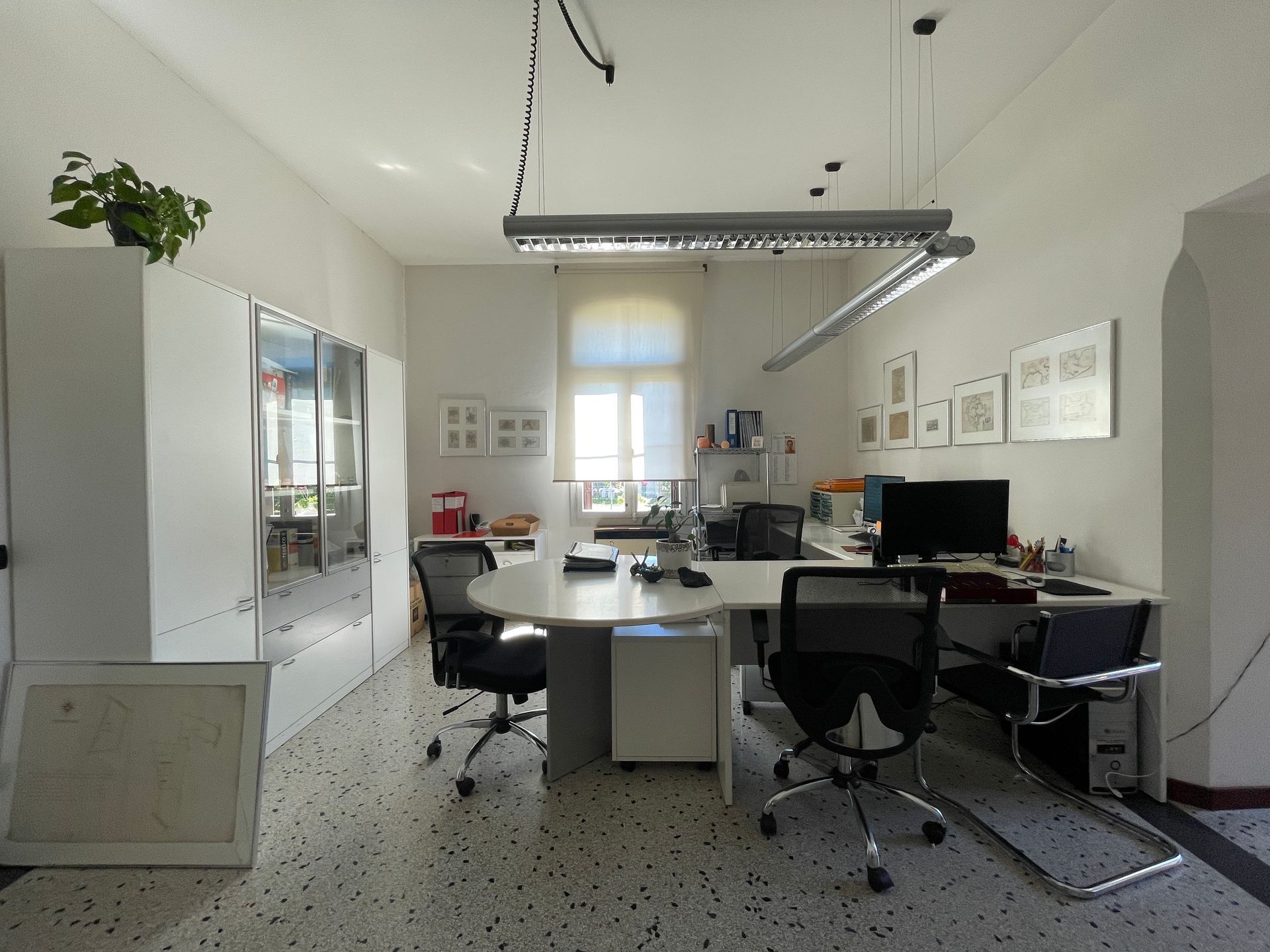 Ufficio moderno con scrivanie, monitor, sedie e un armadio bianco accanto a grandi finestre.