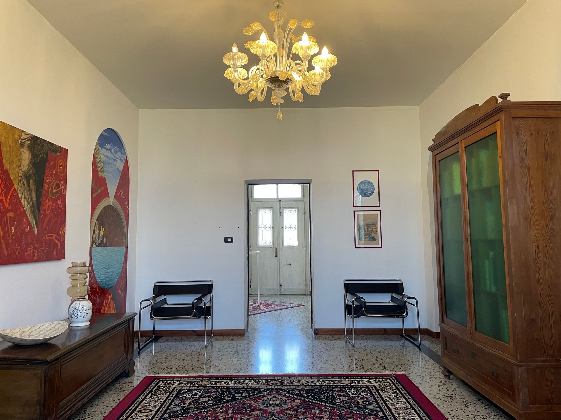 Elegante salotto con lampadario, tappeto persiano, quadri e mobili in legno; porta a vetri centrata sul retro.