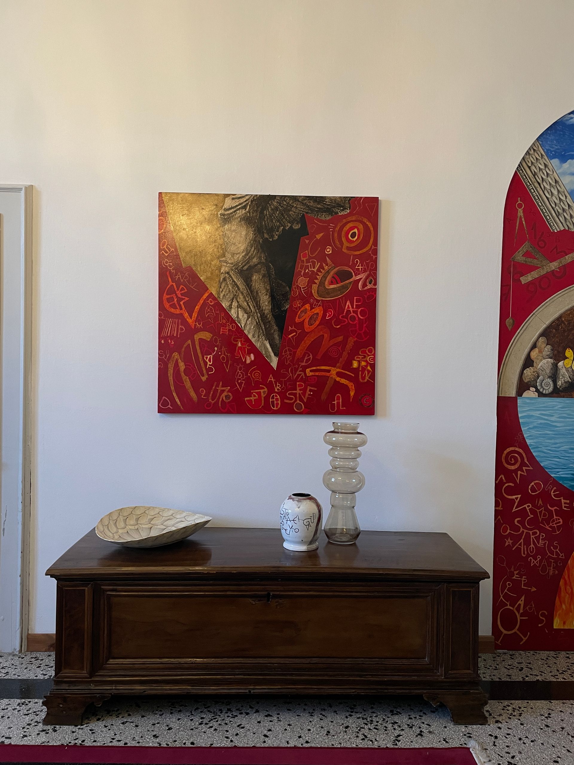 Credenza in legno con oggetti decorativi sotto un quadro astratto rosso e oro in una stanza minimalista