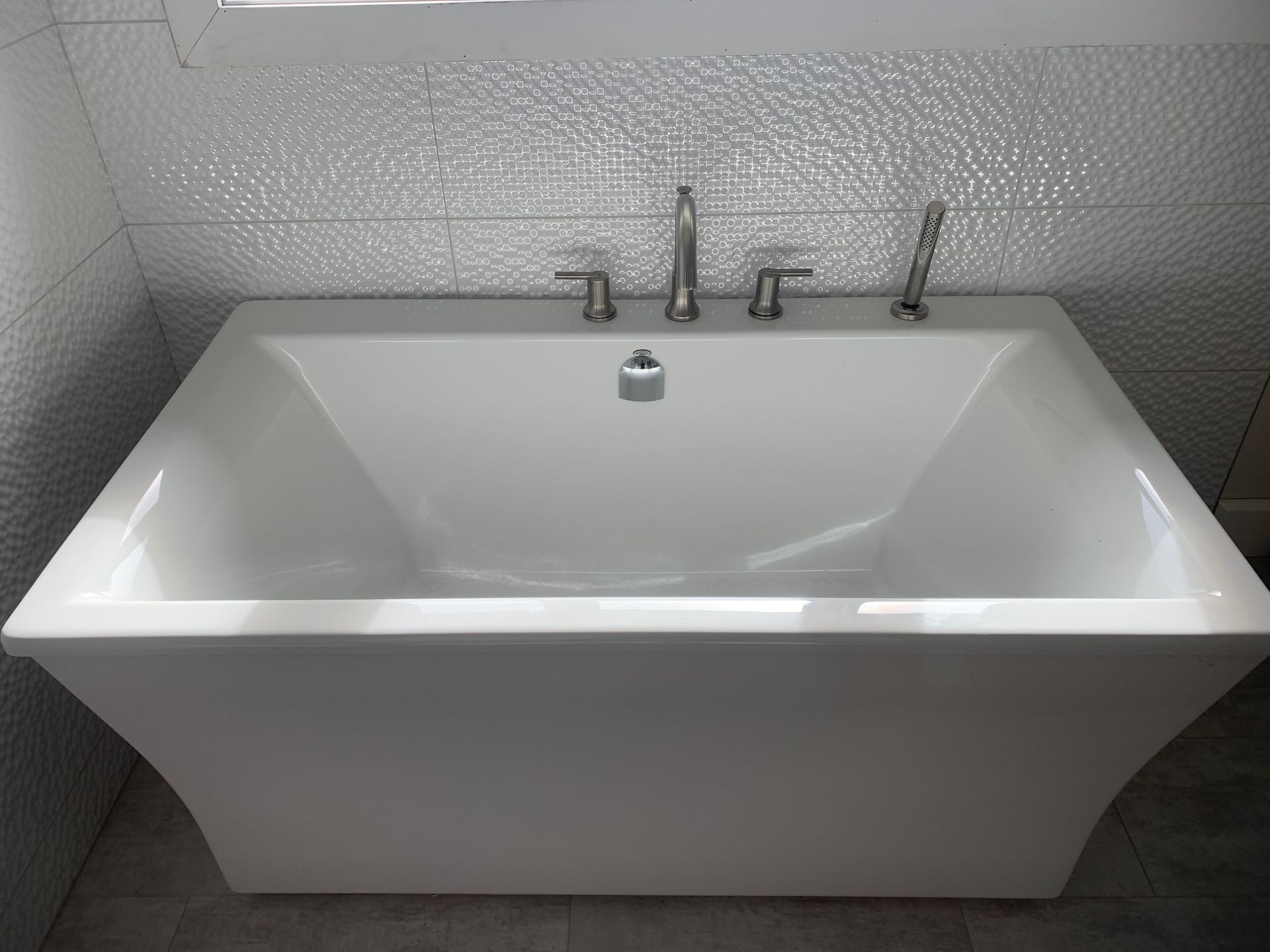 White rectangular bathtub with chrome fixtures set against a textured, light-colored backsplash.