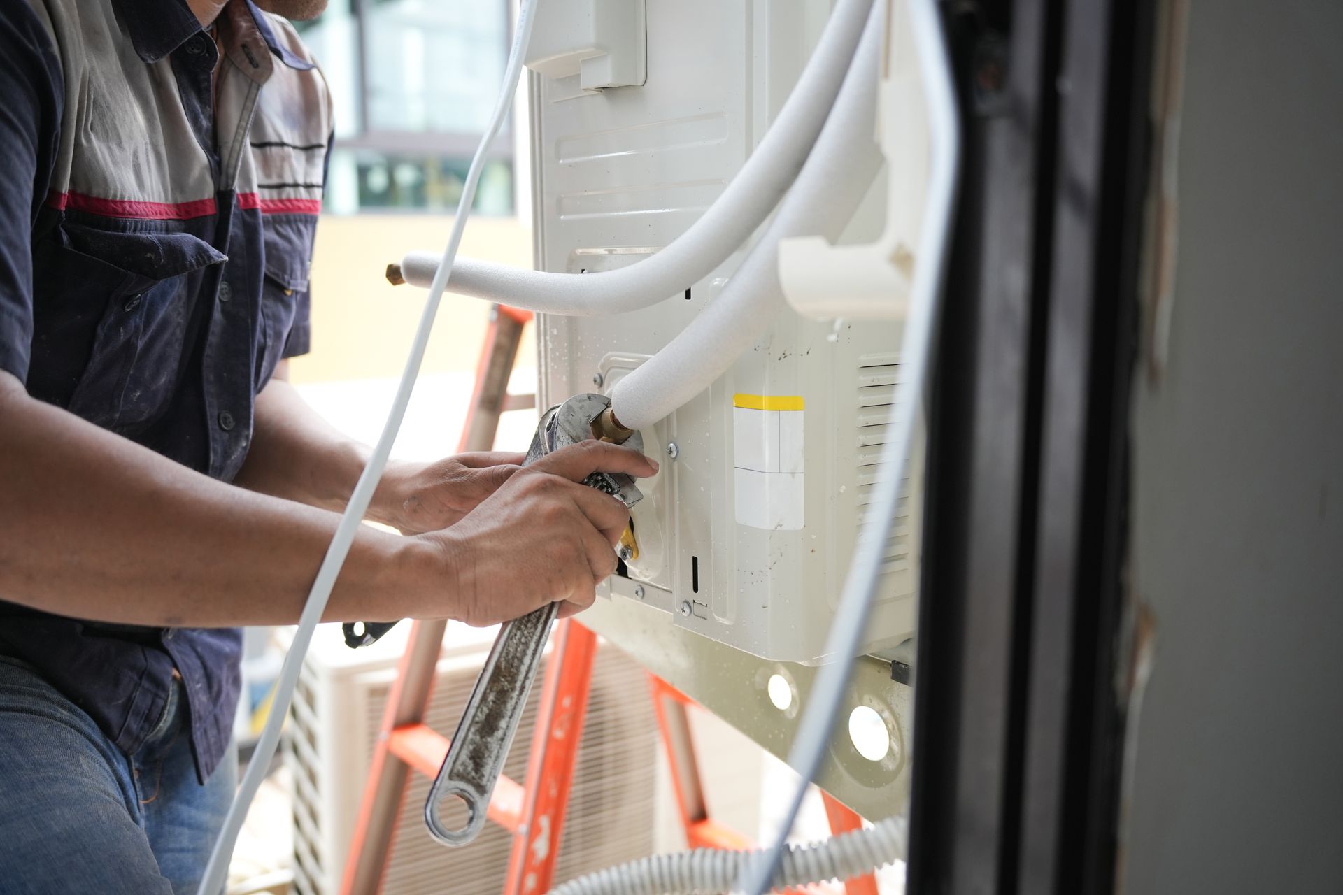Technician connecting wires inside a white HVAC unit on a ladder indoors