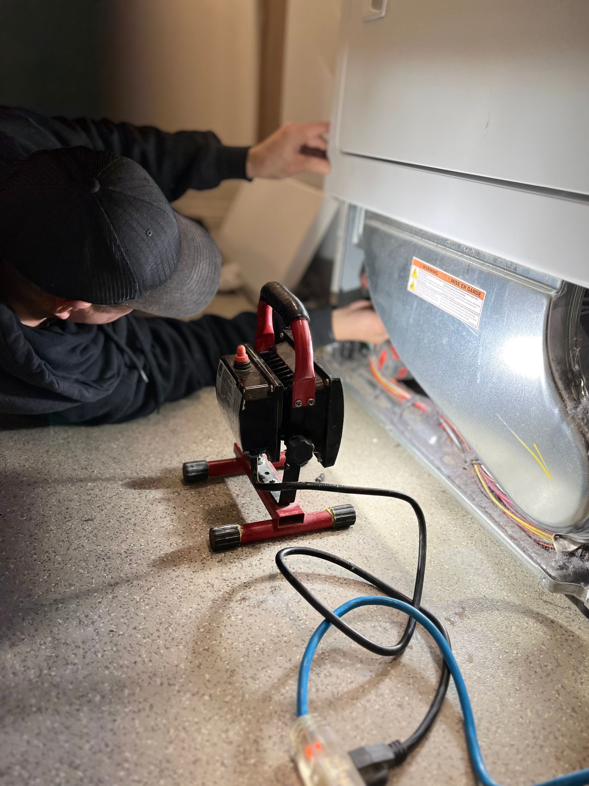 Person repairs appliance, using a portable work light. Gray floor, appliance is silver, person wears black hat and shirt.