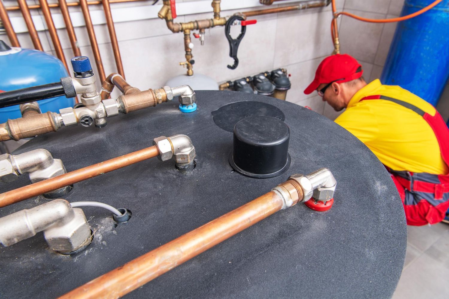Plumber in red cap works on water heater, copper pipes, indoor setting.