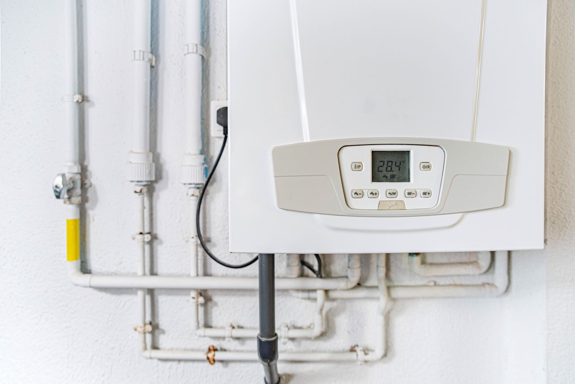 A white boiler is hanging on a white wall next to pipes.