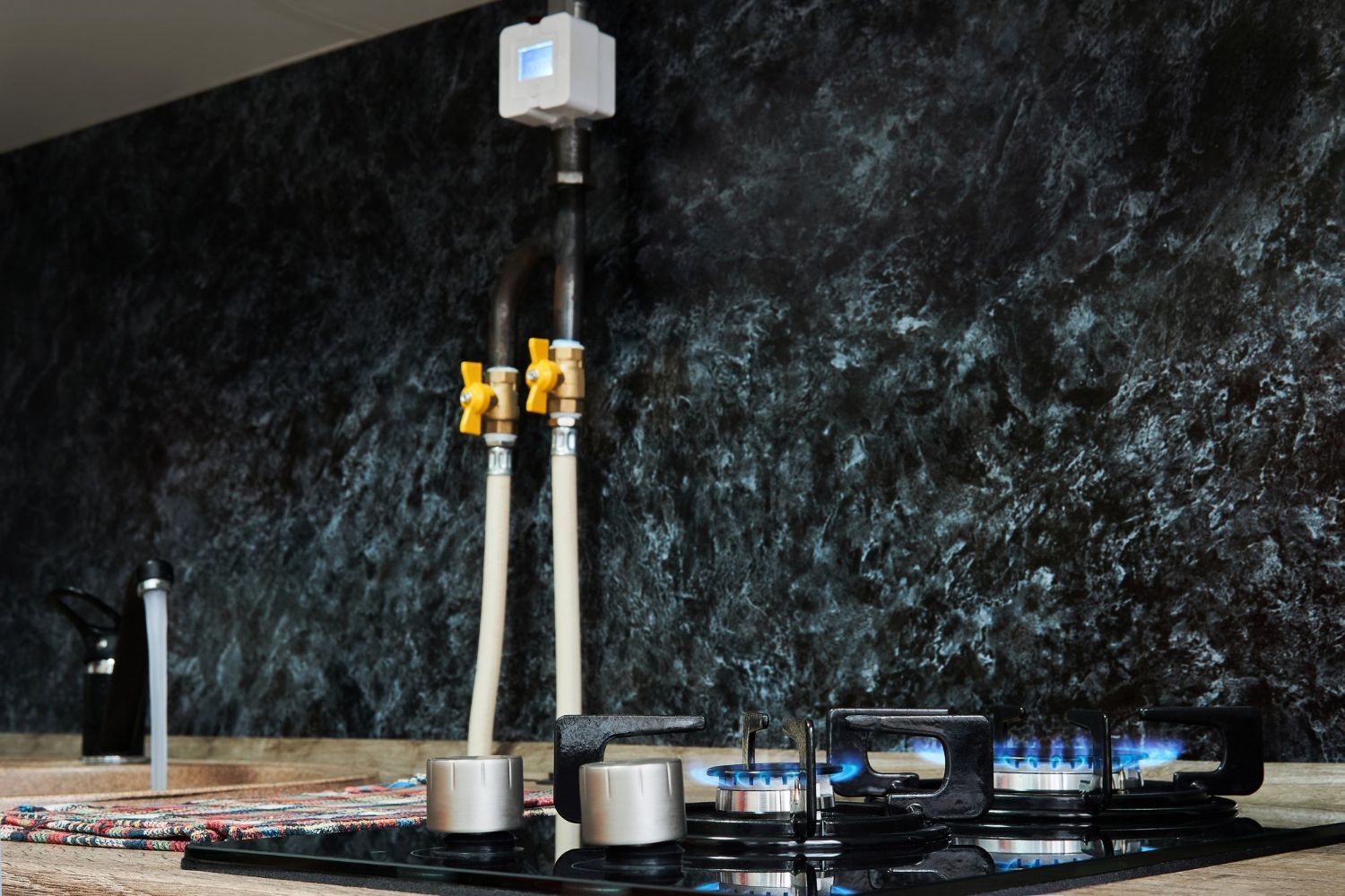 Gas stove with blue flames, gas pipes, and yellow valves against a dark textured wall.
