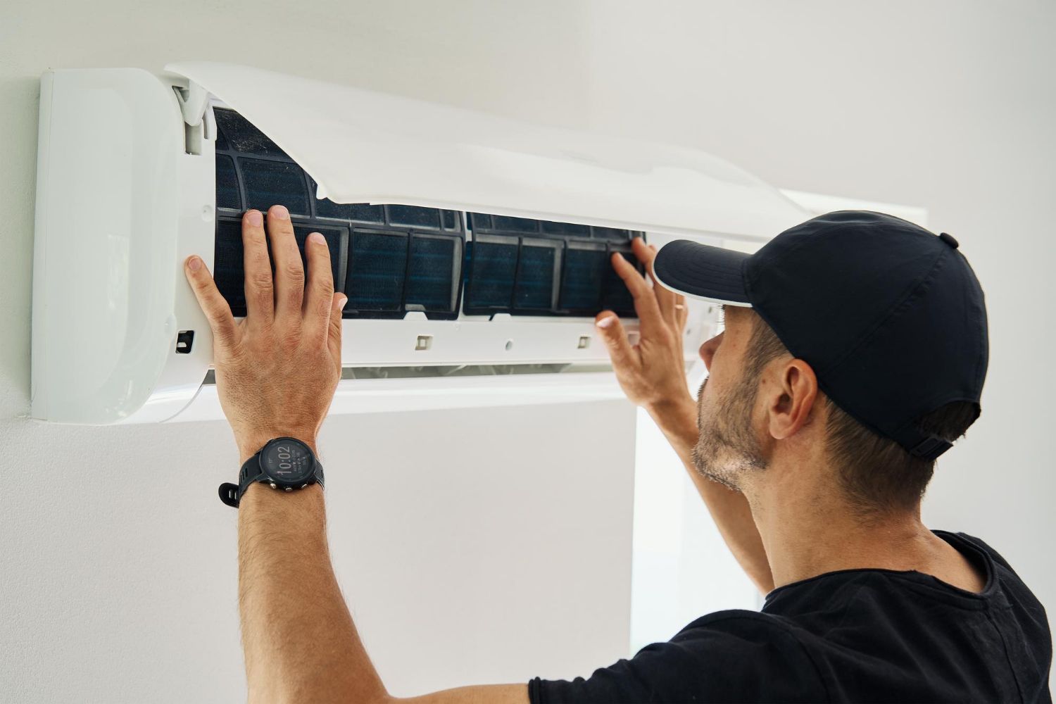 Man in black cap servicing a white wall-mounted air conditioner, removing a filter.