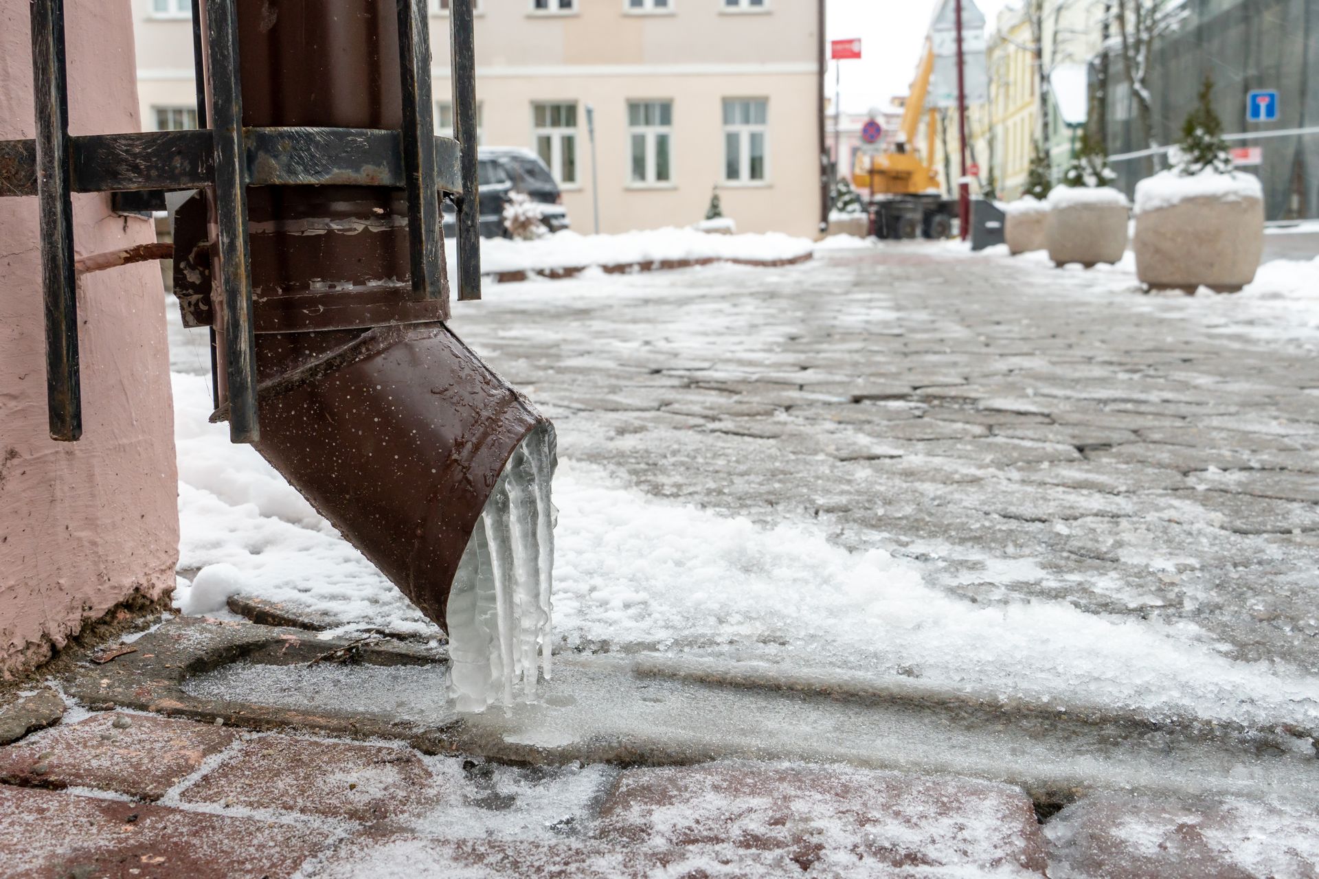 Brown gutter with icicles, sidewalk covered in snow, buildings in background.