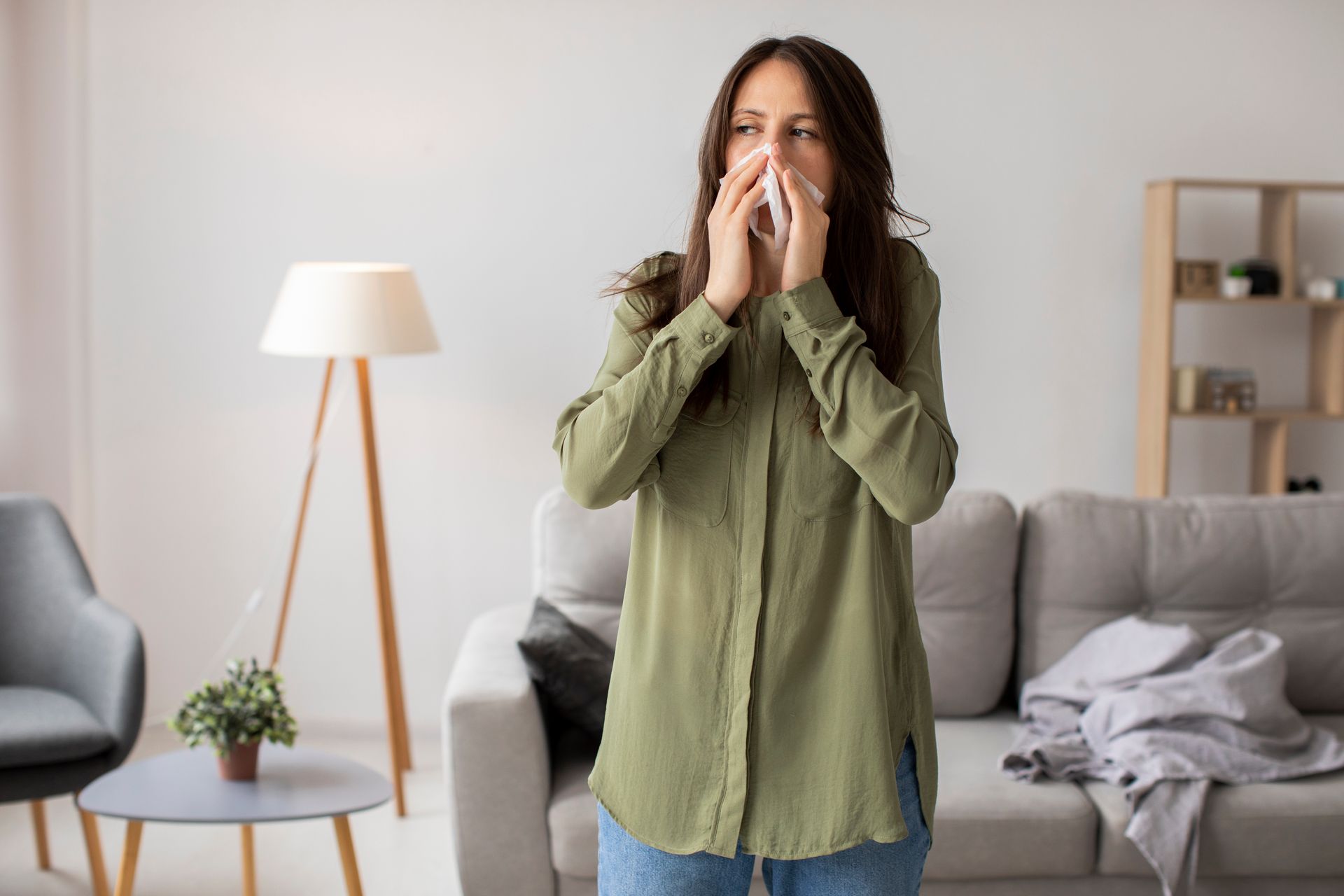 Woman in green shirt and jeans blowing her nose indoors.