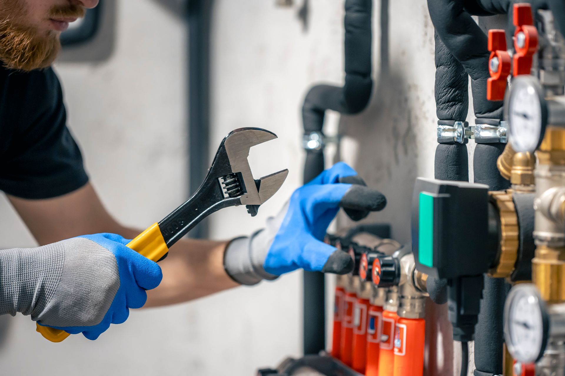 Plumber with wrench, fixing pipes in a mechanical room, wearing gloves.
