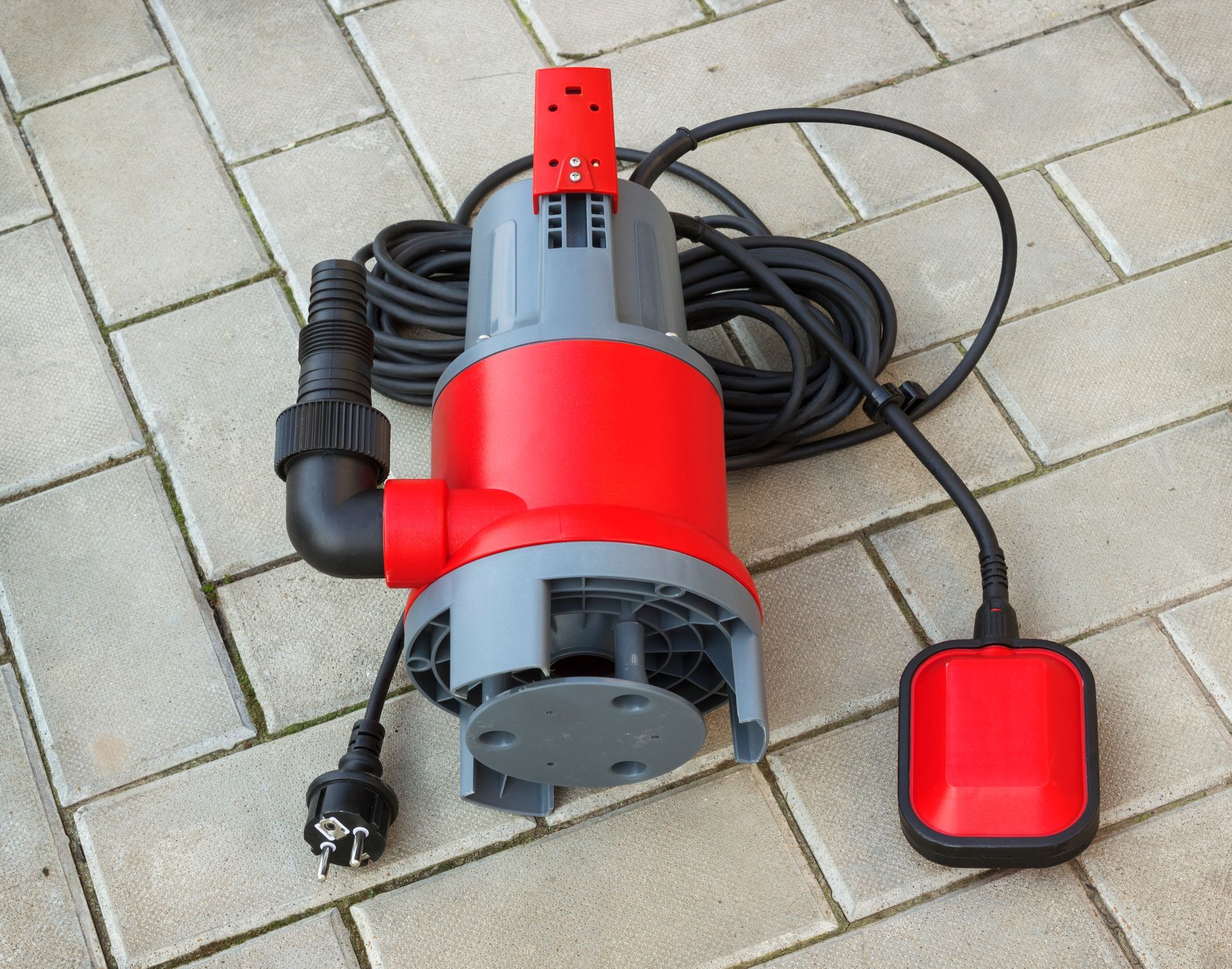 Red and gray submersible water pump with black hose and float switch, on brick patio.