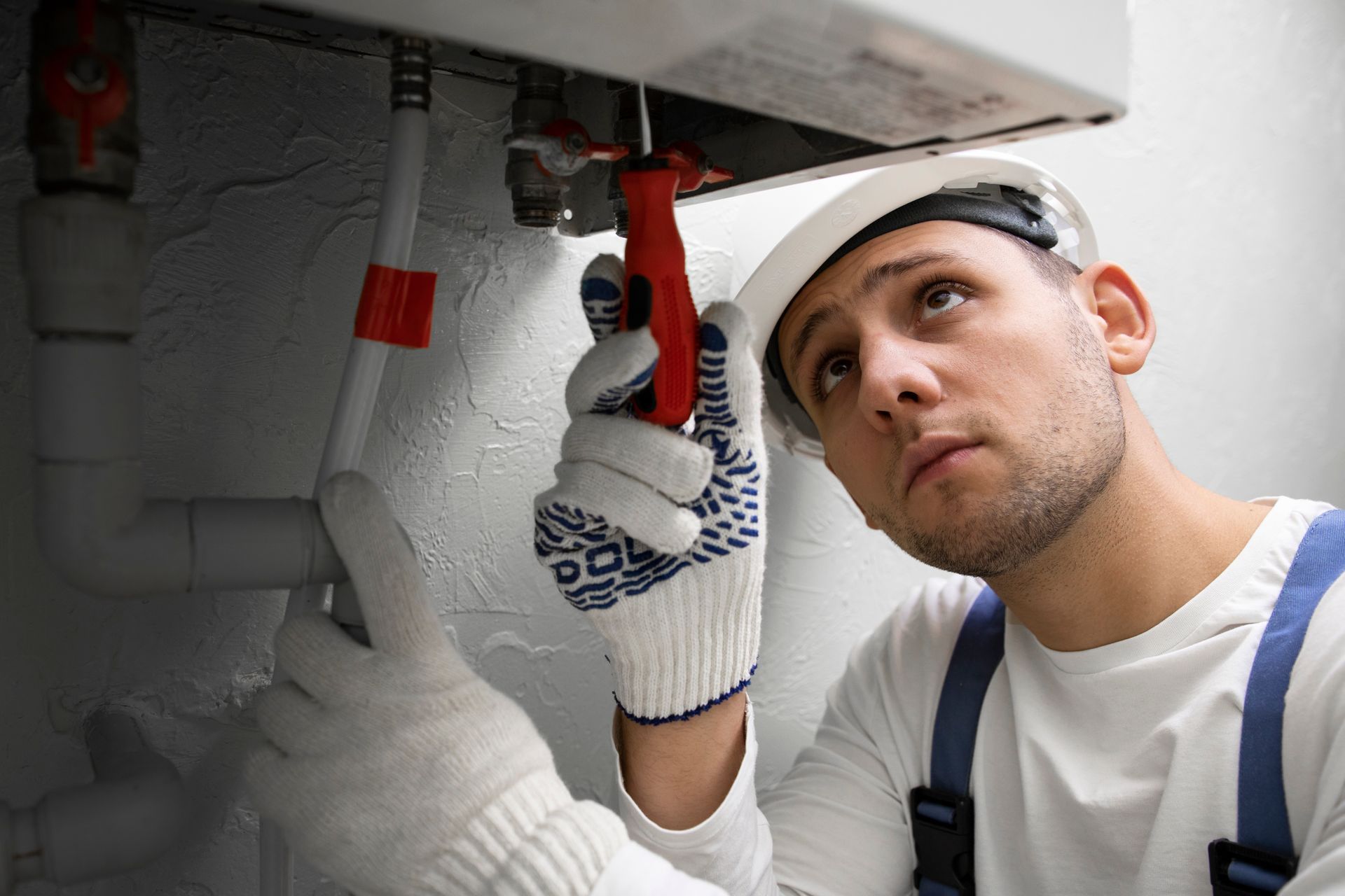 A plumber in a hard hat fixes pipes with a wrench, indoors.