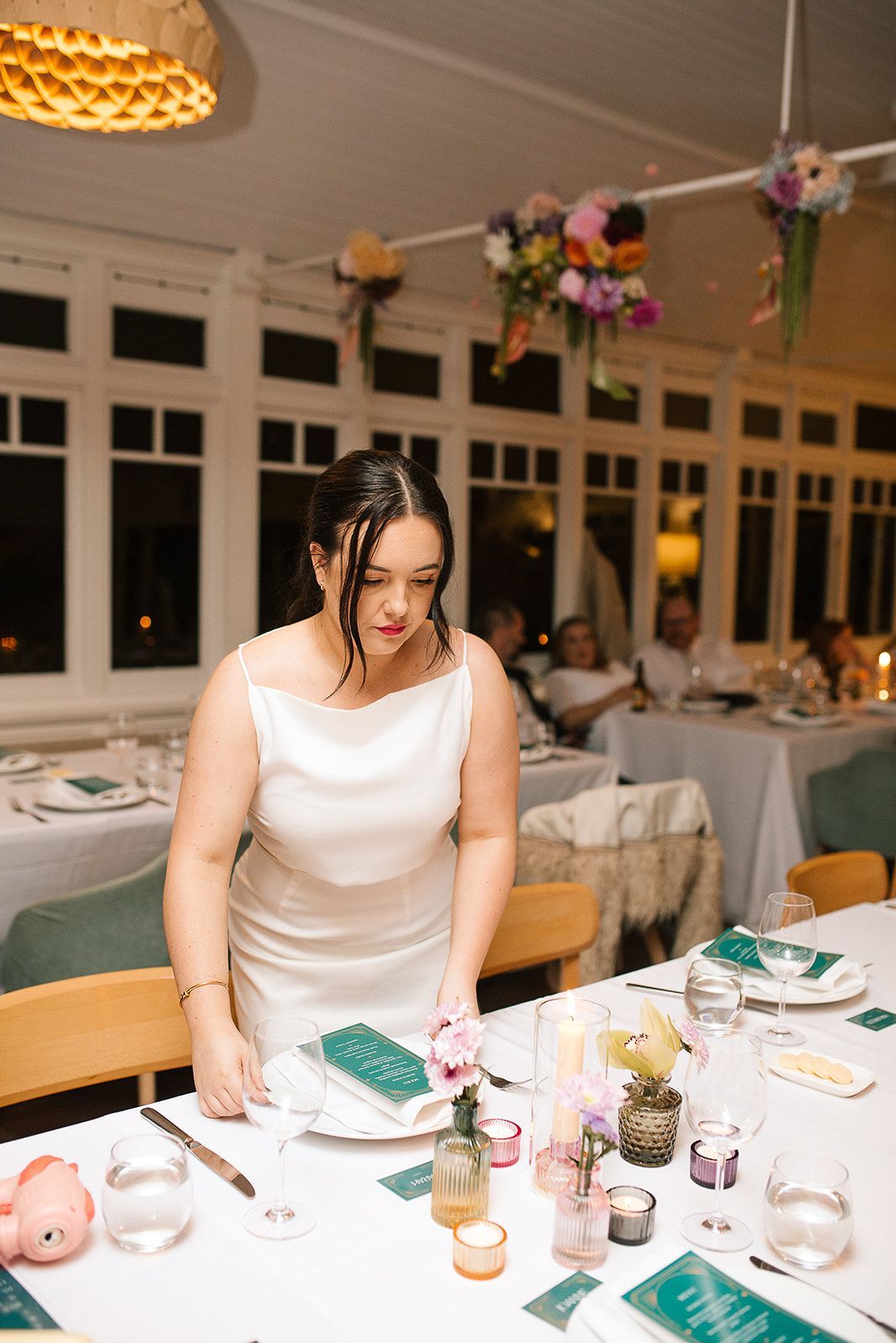 Woman setting a table at a wedding reception with floral decorations; indoor setting with windows and guests.