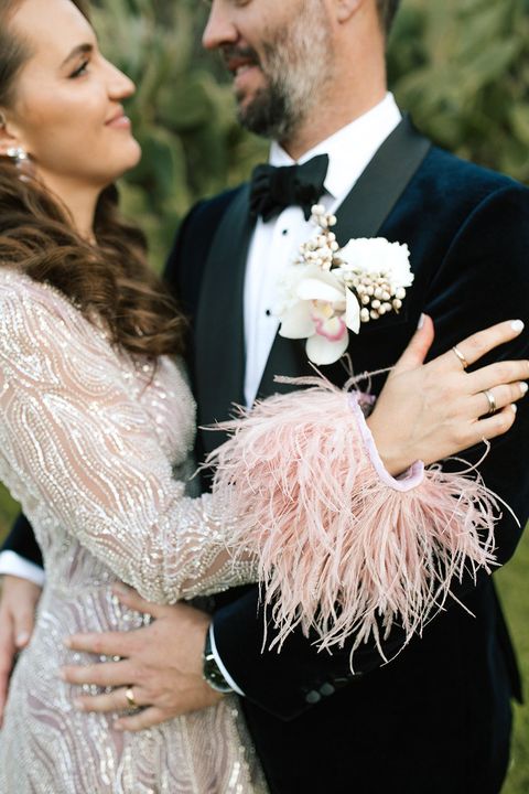 Bride and groom embrace; she wears a shimmering gown, he wears a tuxedo. Pink feather cuffs on his arm.