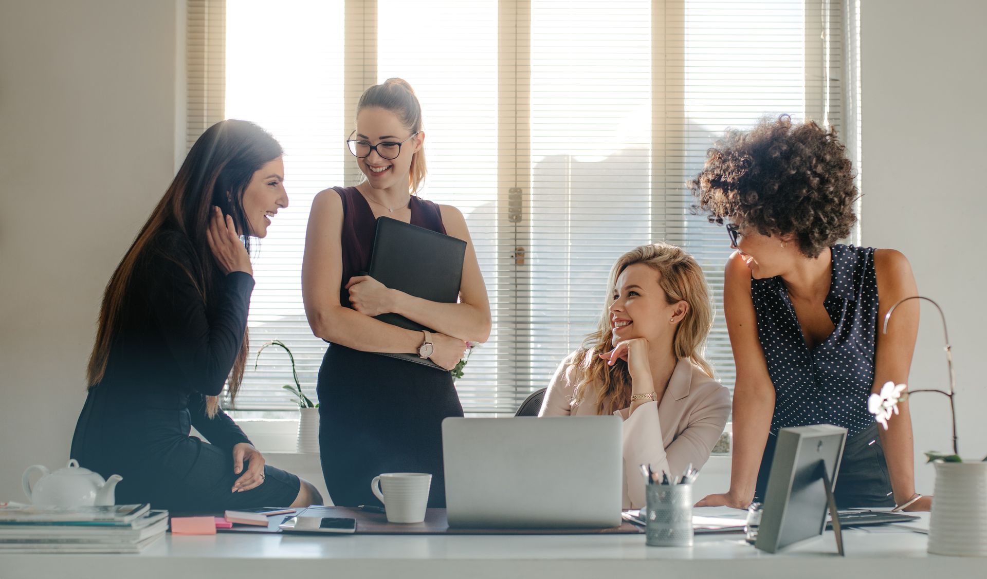 Four women in an office setting, talking and smiling near a laptop, window with sunlight.
