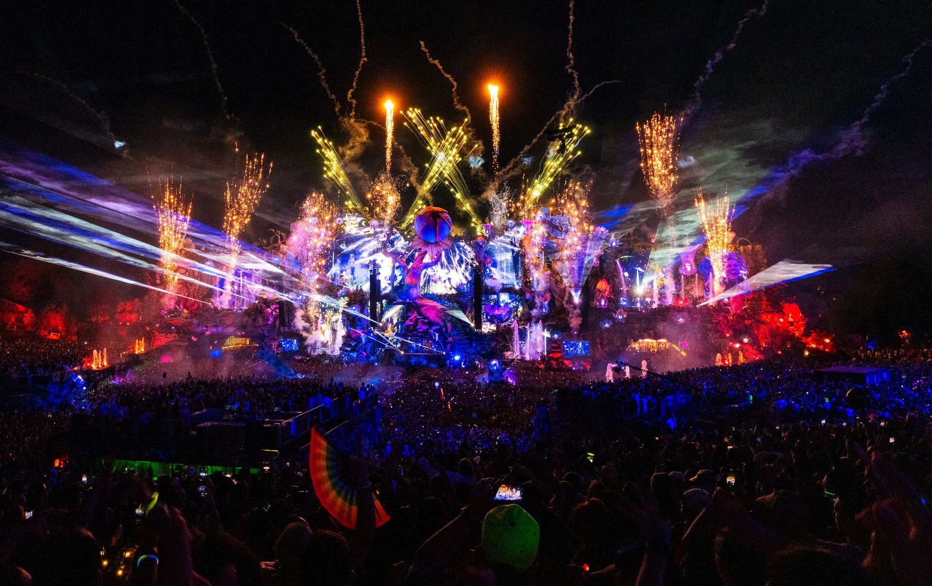 Crowded nighttime concert with bright stage lights, fireworks, and a cheering crowd