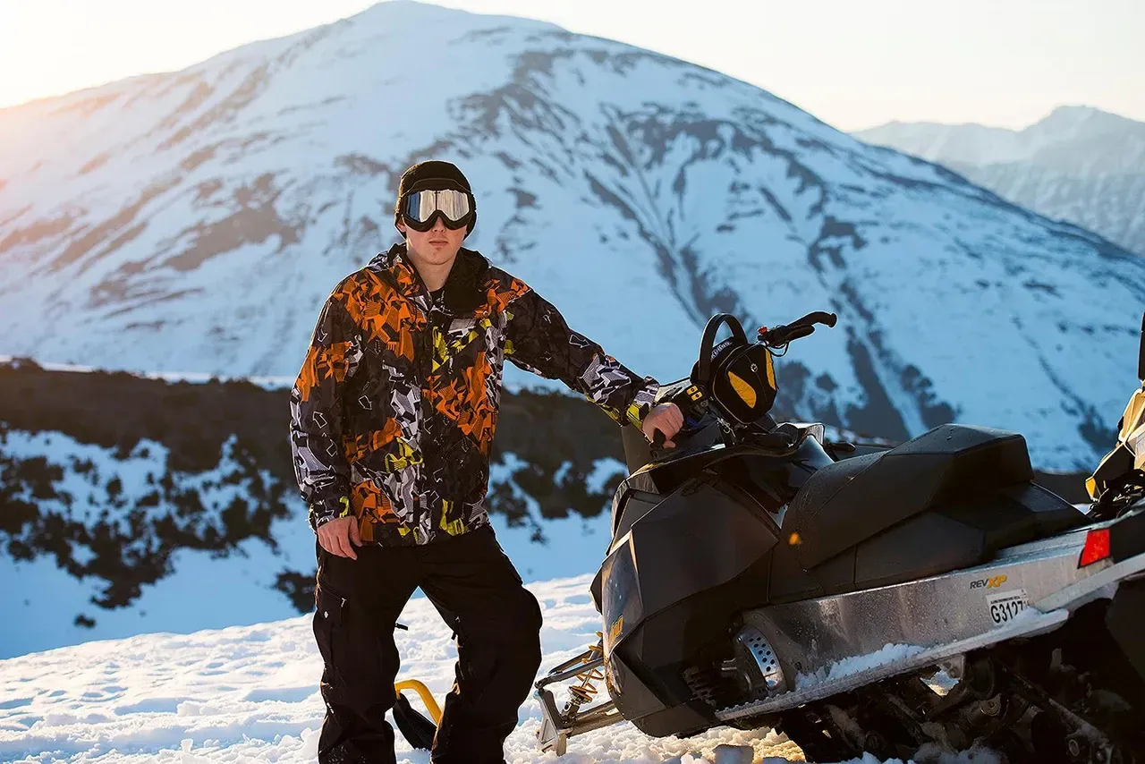 Person standing beside a snowmobile in snowy mountains at sunset