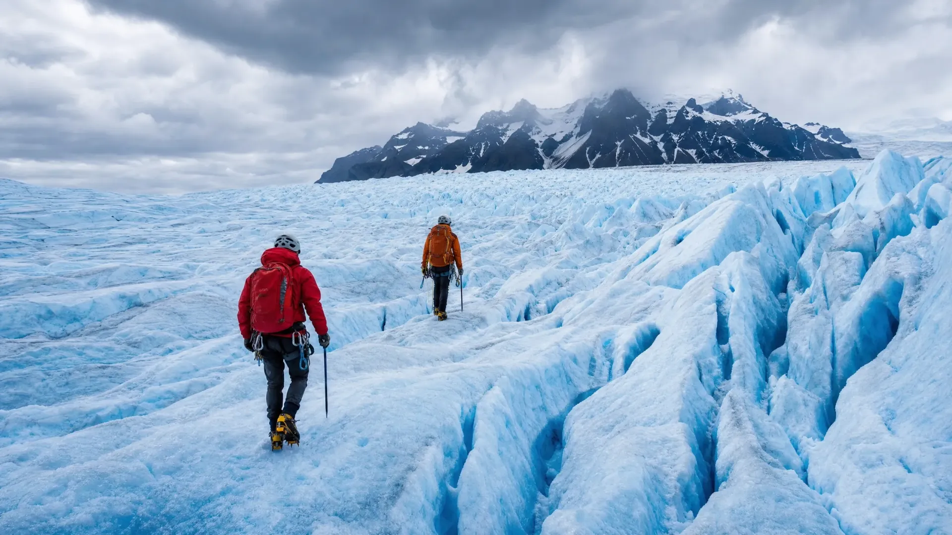 Two hikers in red and orange jackets trek across a blue glacier toward snowy mountains.