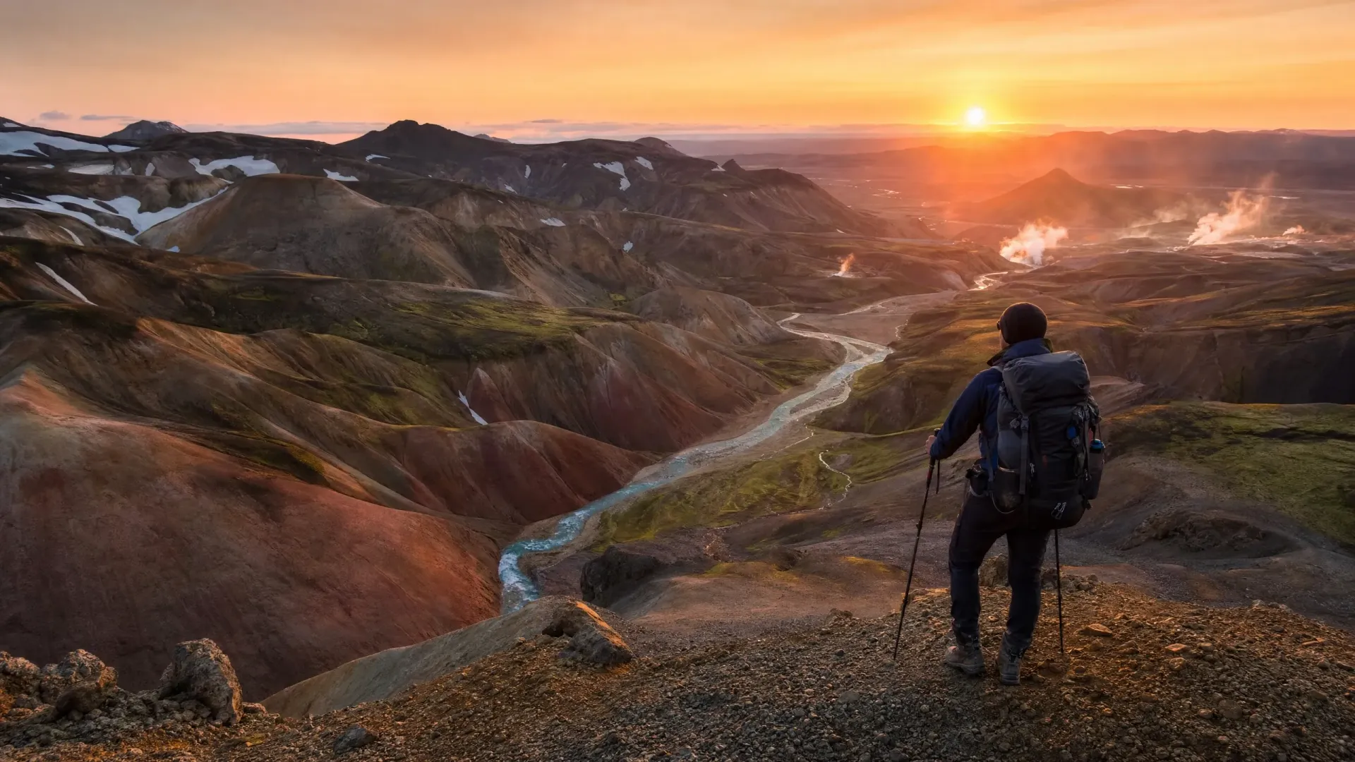 Hiker overlooking a rugged valley at sunrise, with a winding stream and glowing orange sky.