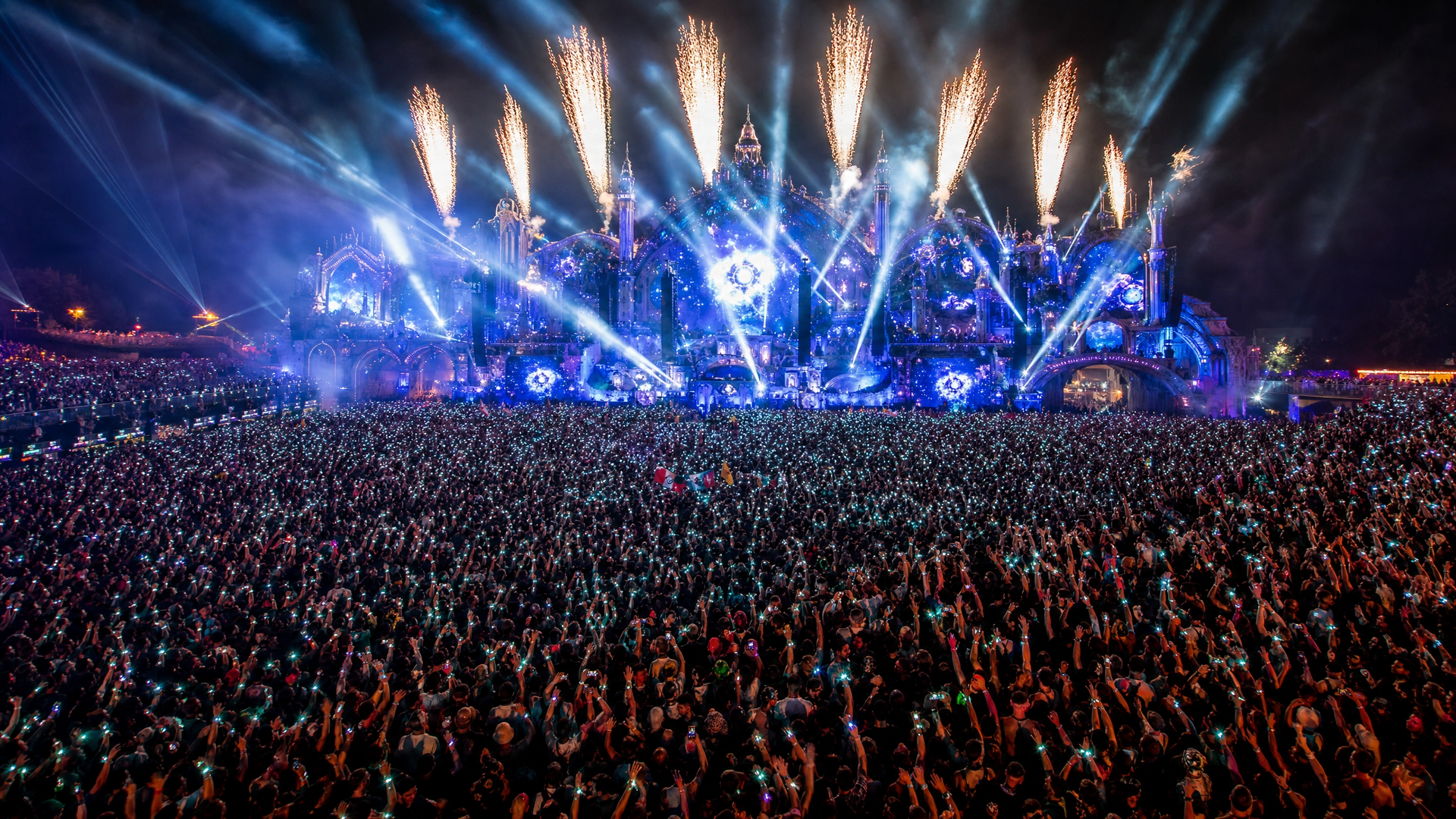 Night concert crowd facing a blue-lit stage with fireworks and bright pyrotechnics overhead