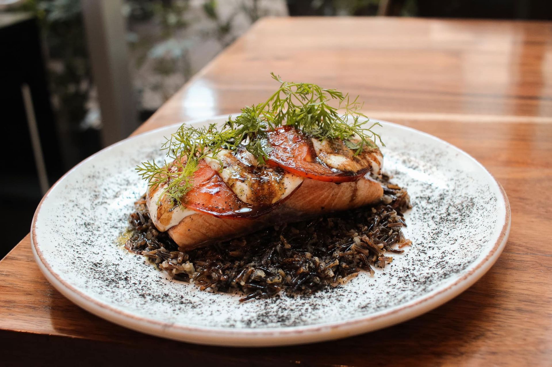 Salmón sellado en un plato blanco con hierbas y salsa oscura sobre una mesa de madera.
