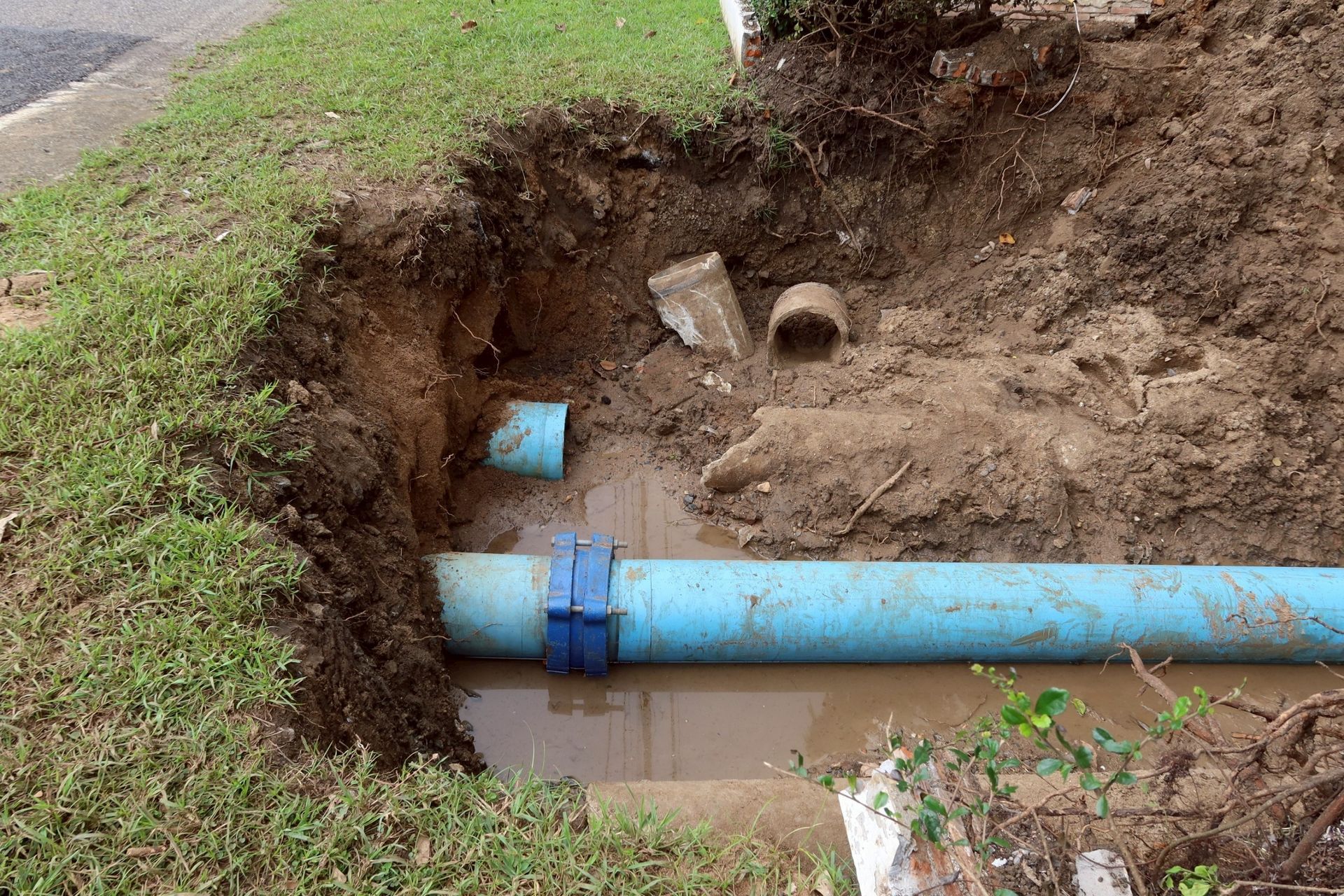 Excavation exposing blue water pipe with a coupling, in a muddy yard next to grass.