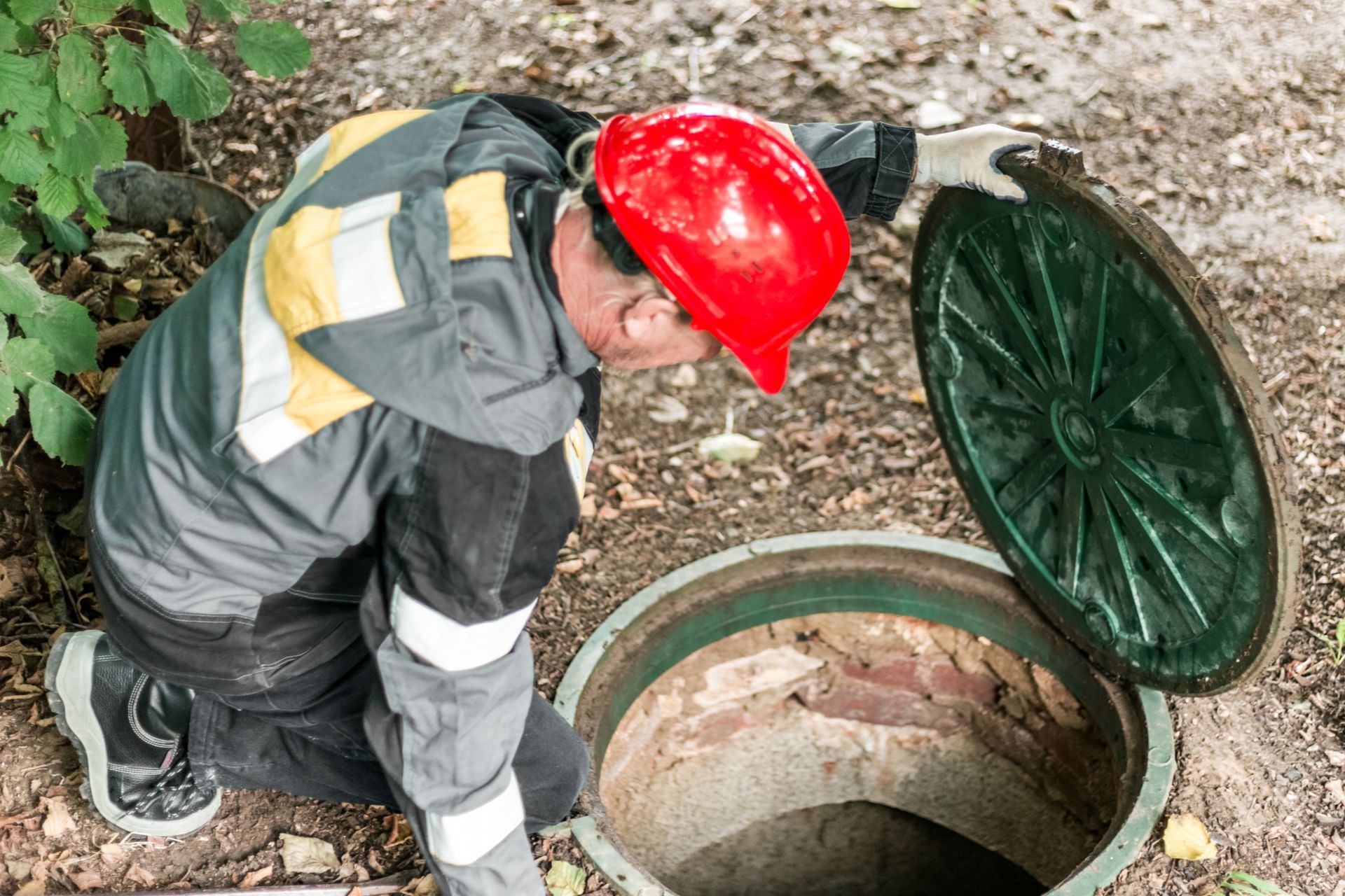 Man in hard hat examines an open sewer manhole in a grassy area.