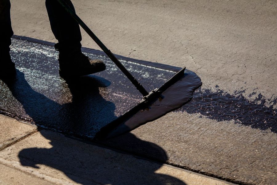 A worker uses a squeegee to spread black asphalt sealant across a section of pavement.