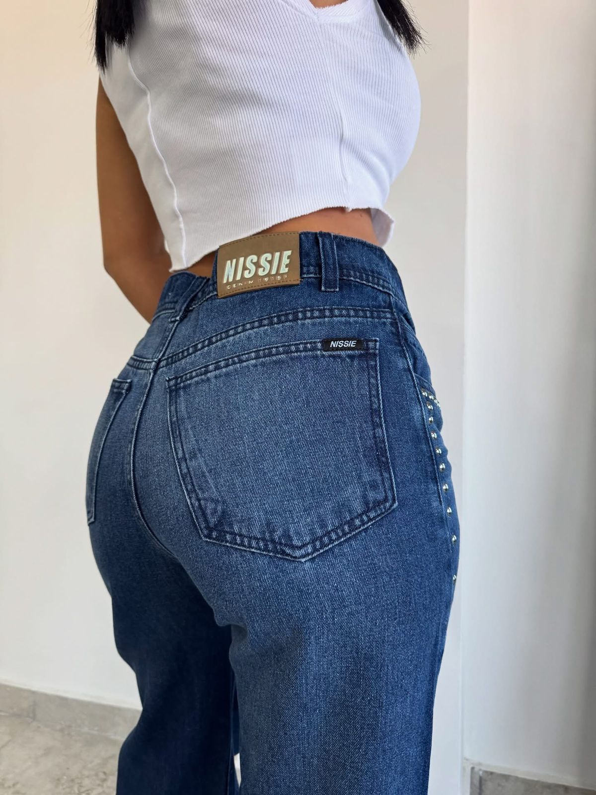 Una mujer que viste un top blanco y jeans anchos está apoyada contra una pared.