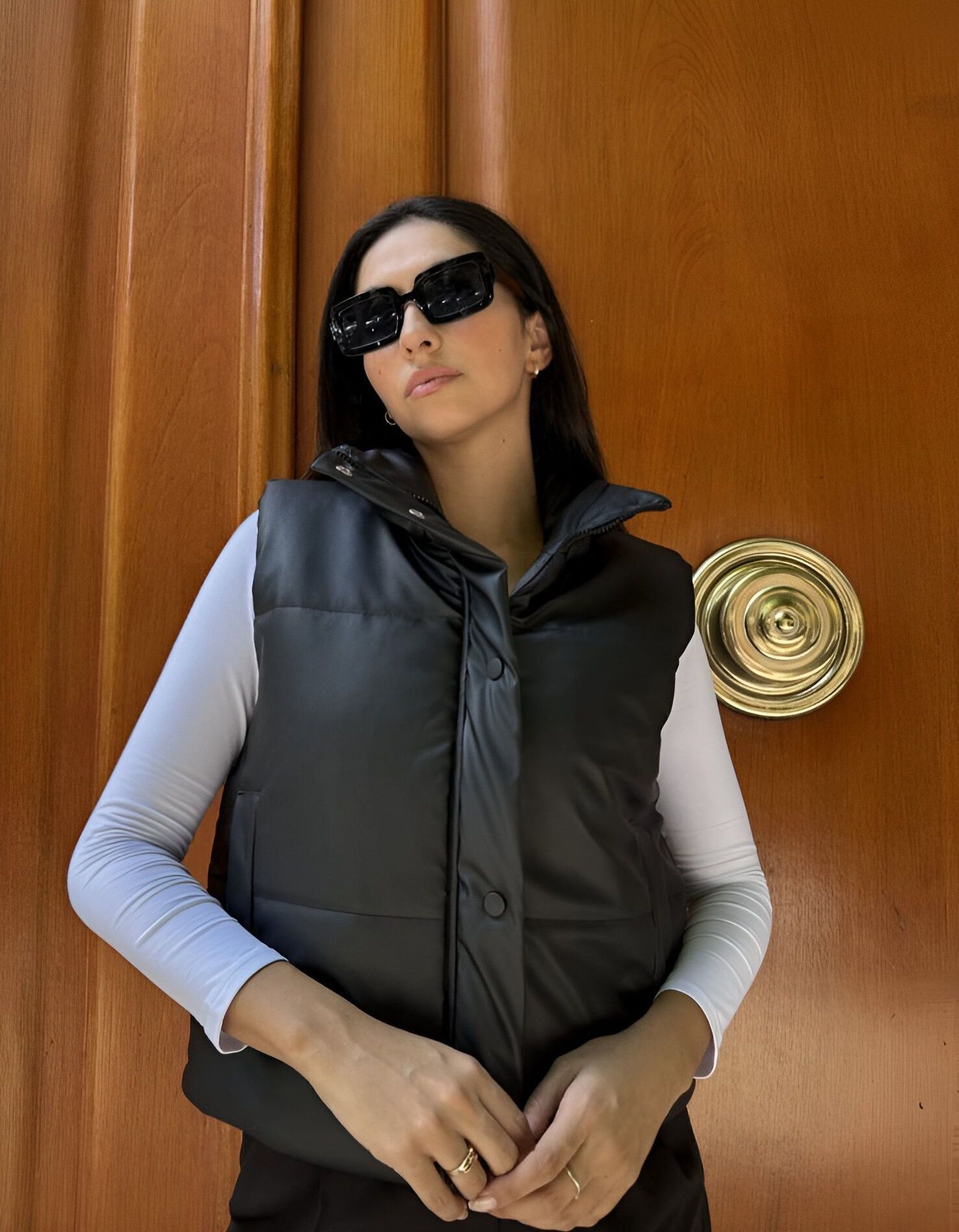 Una mujer que lleva gafas de sol y un chaleco negro está parada frente a una puerta de madera.