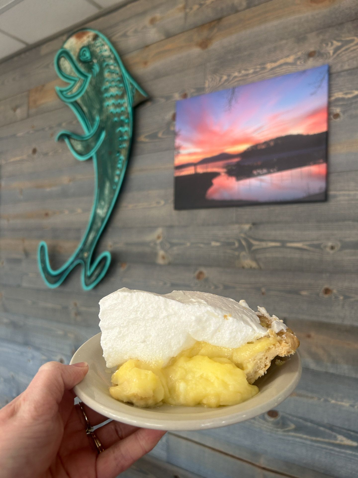 Person holding a plate with a slice of pie in front of a wall with a fish sculpture and landscape photo.