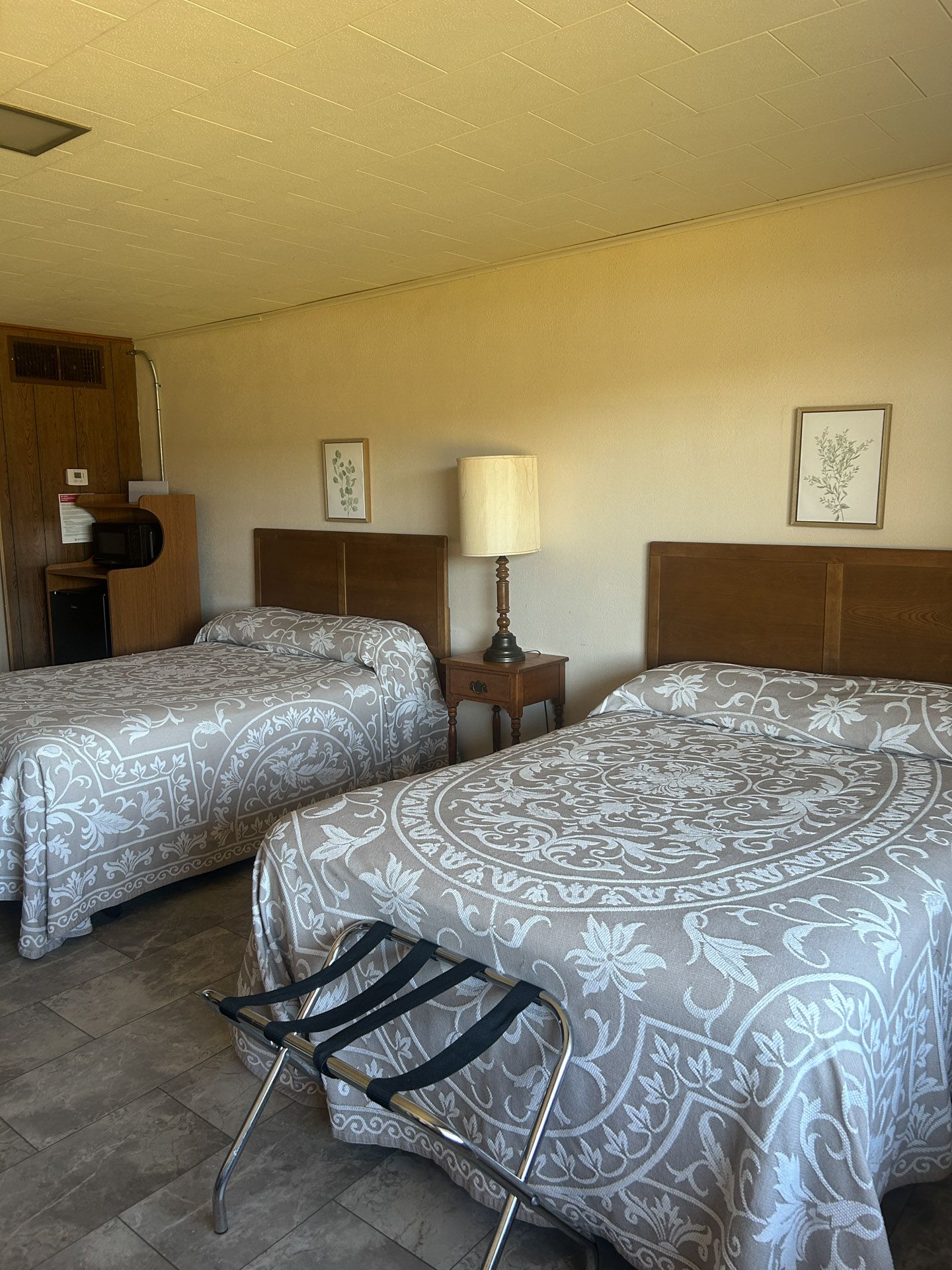 Two beds in a motel room with floral patterned bedspreads, lamp, and luggage rack.