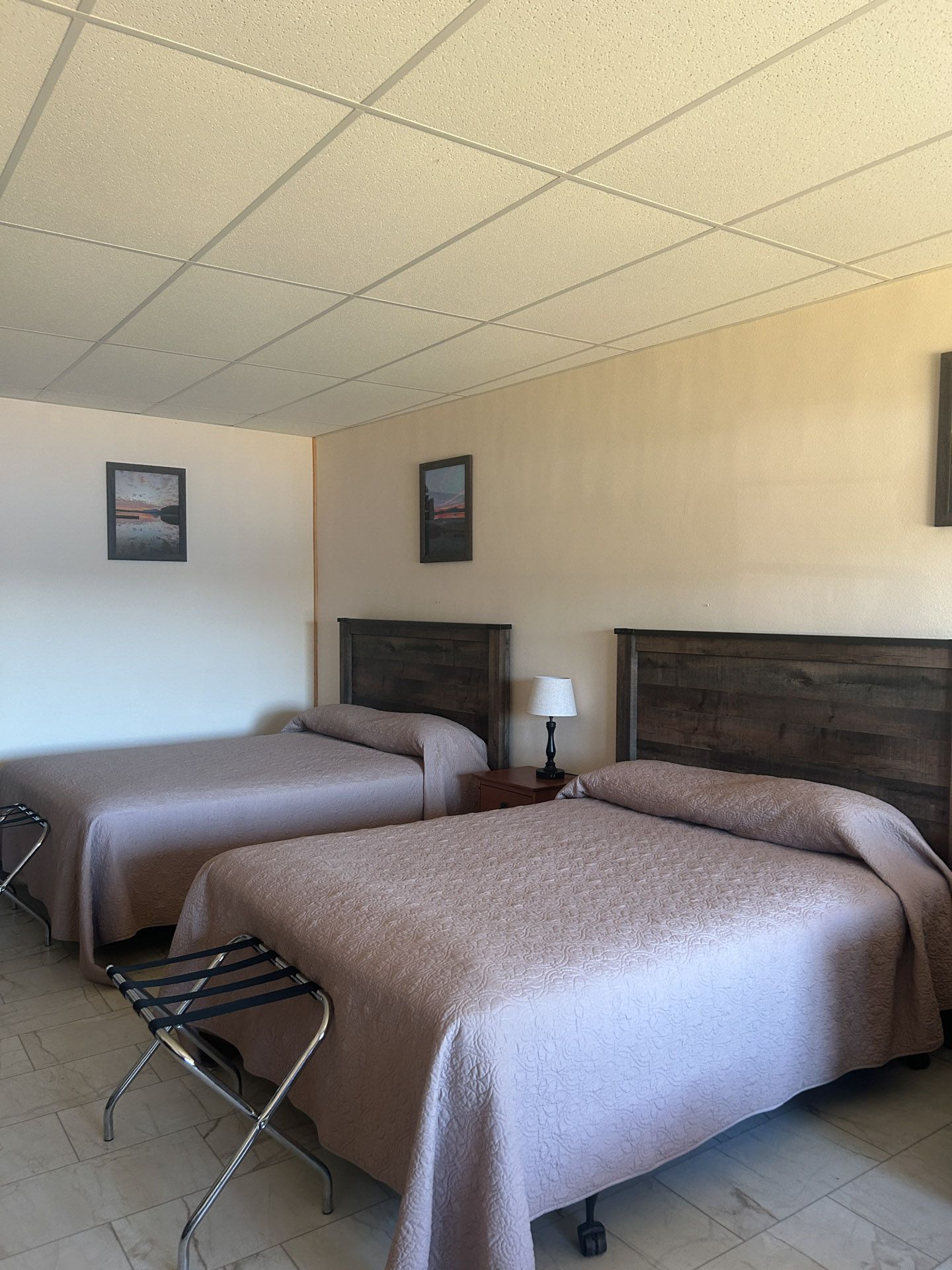 Two beds in a motel room with a nightstand and two framed pictures on the wall.