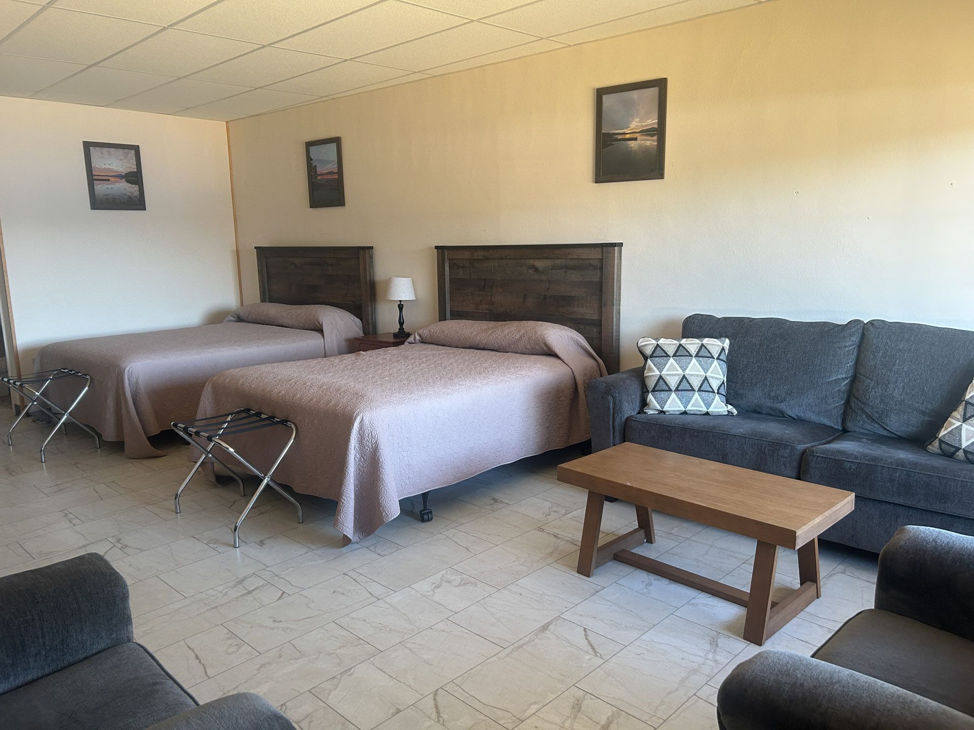 Hotel room with two beds, a couch, armchairs, small table, and framed art. Beige walls, tiled floor.