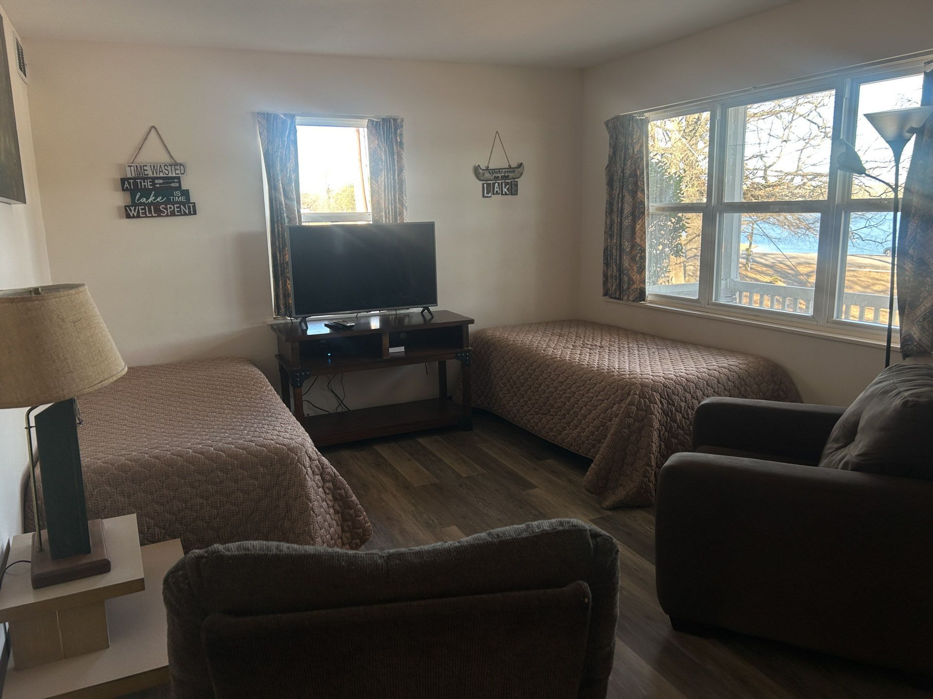 Living room with two beds, TV, armchair, and window overlooking a body of water.