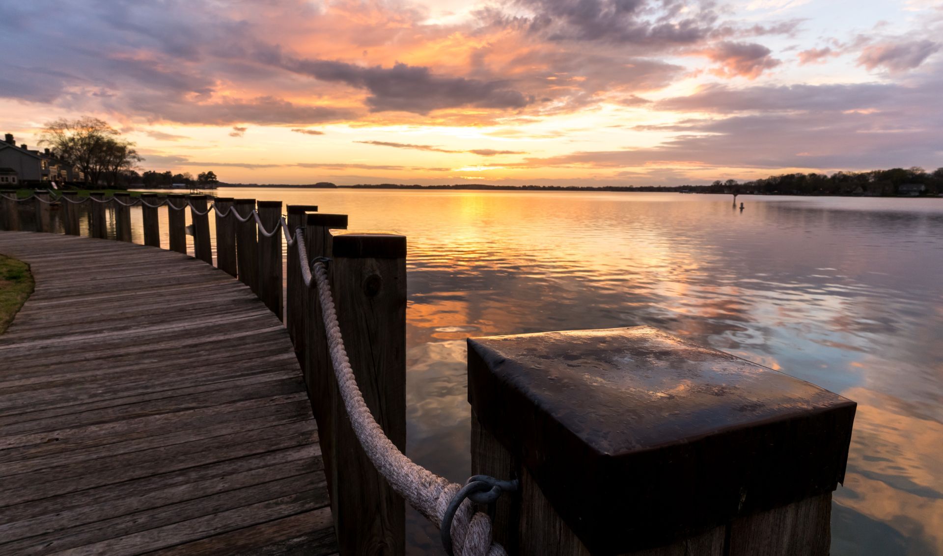 Wooden boardwalk leading to a calm lake reflecting a vibrant sunset with orange and purple hues.