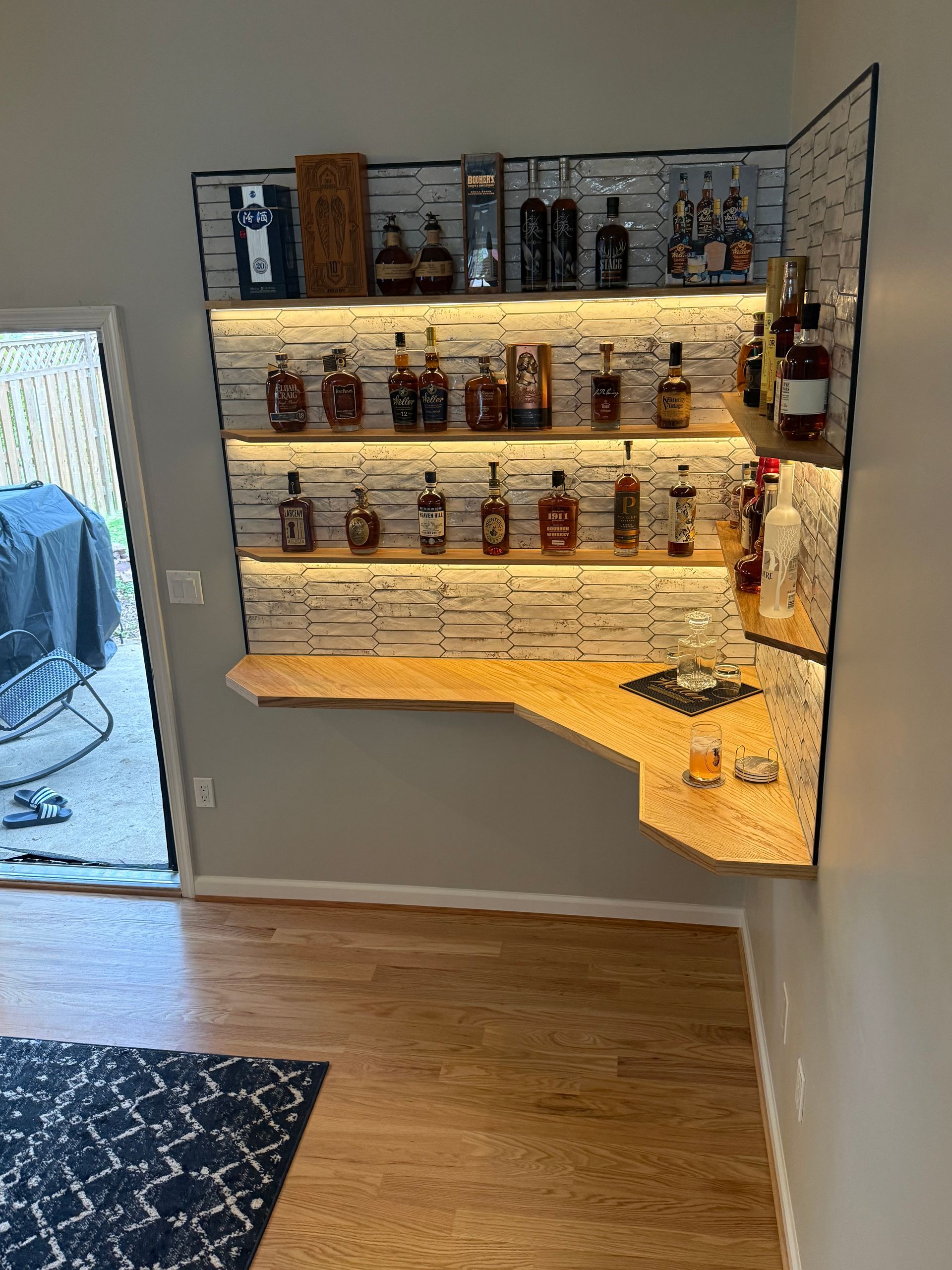 Wall-mounted corner bar shelf with illuminated liquor bottles in a modern room, beside a doorway and rug