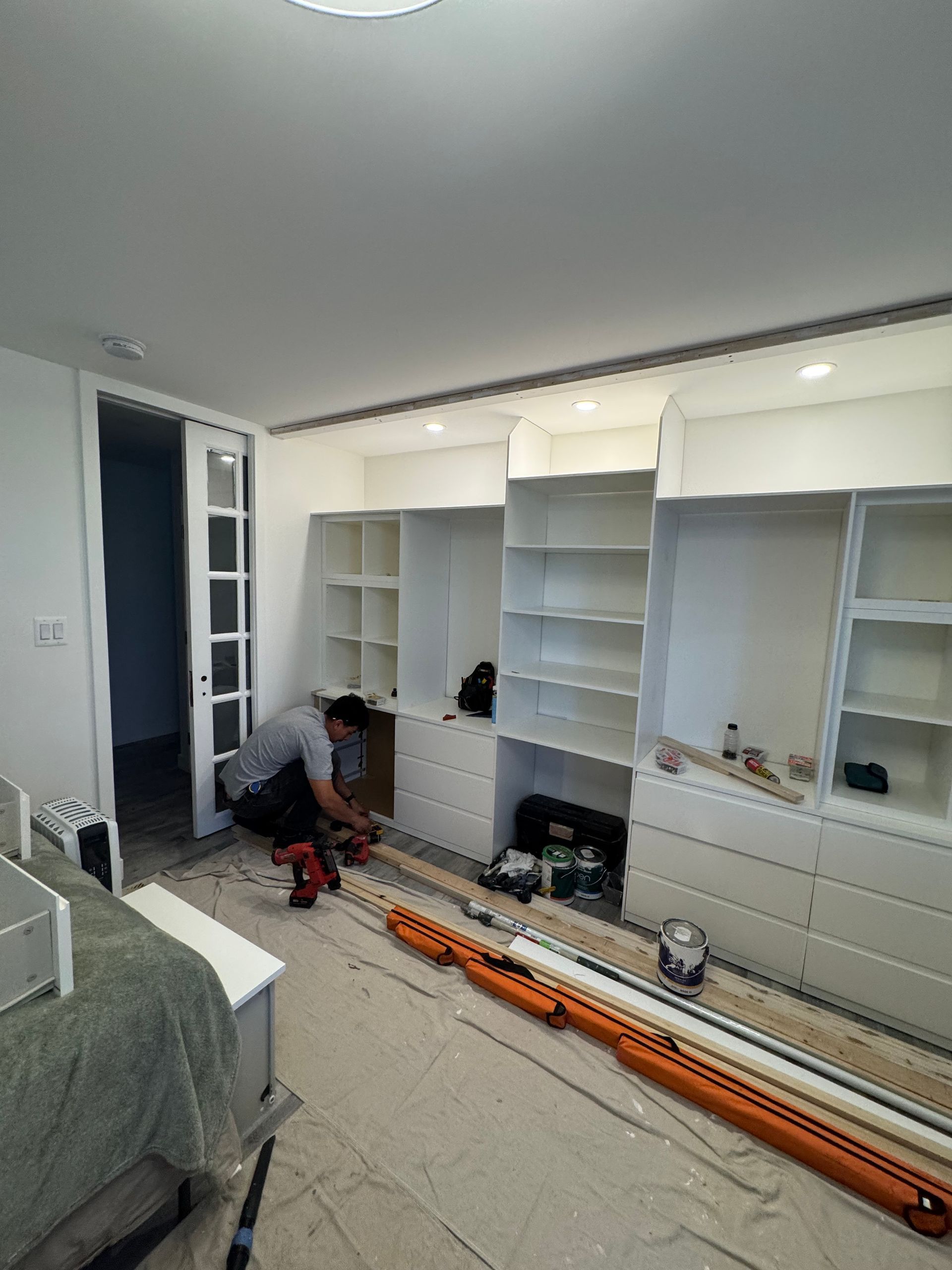 Person assembling white built-in shelves in a bright room with tools on the floor.