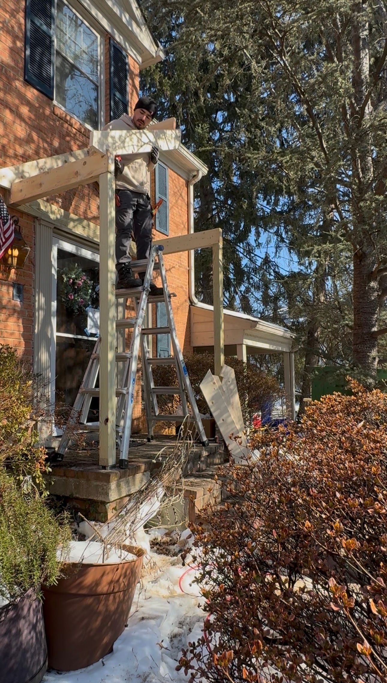 Snowy porch with a ladder leaned against a brick house beside bare trees and a bush