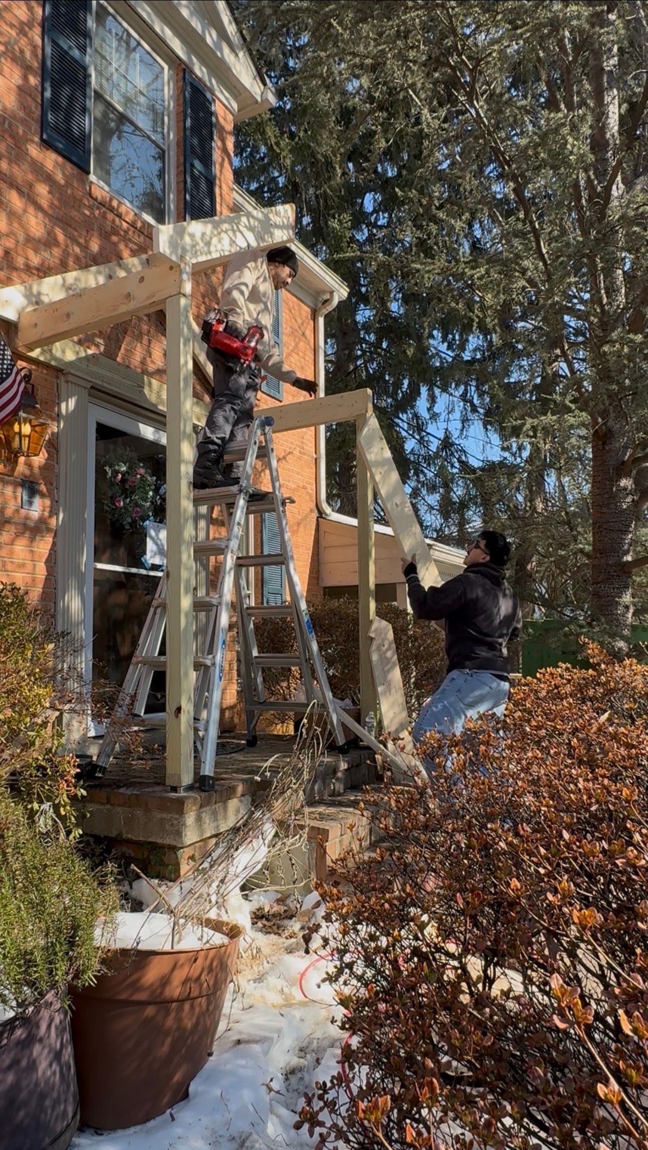 Person on a ladder decorating a brick house with red holiday ornaments in a snowy yard