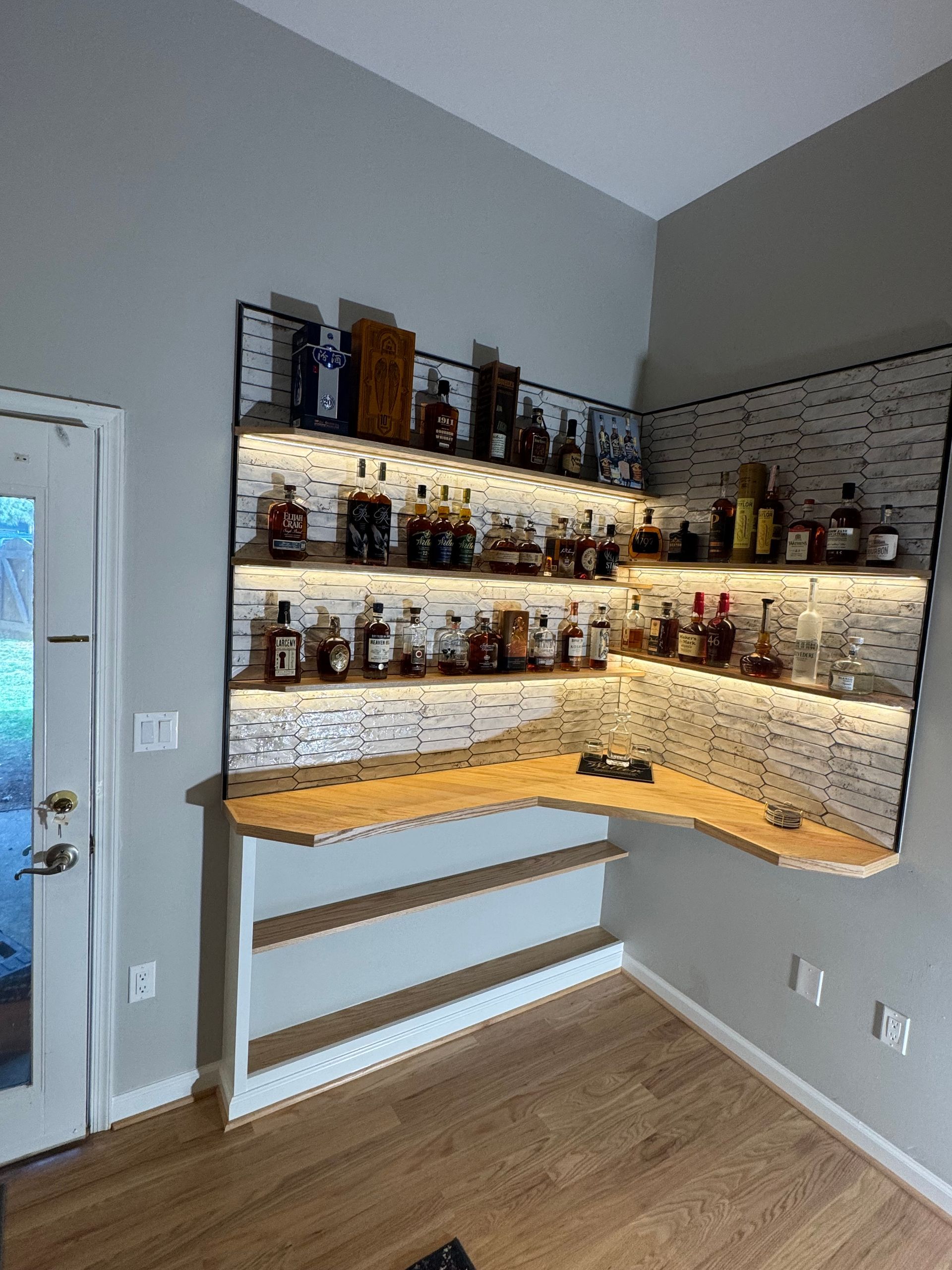 Corner bar shelves with bottles, warm under-shelf lighting, and a wooden counter in a gray room.