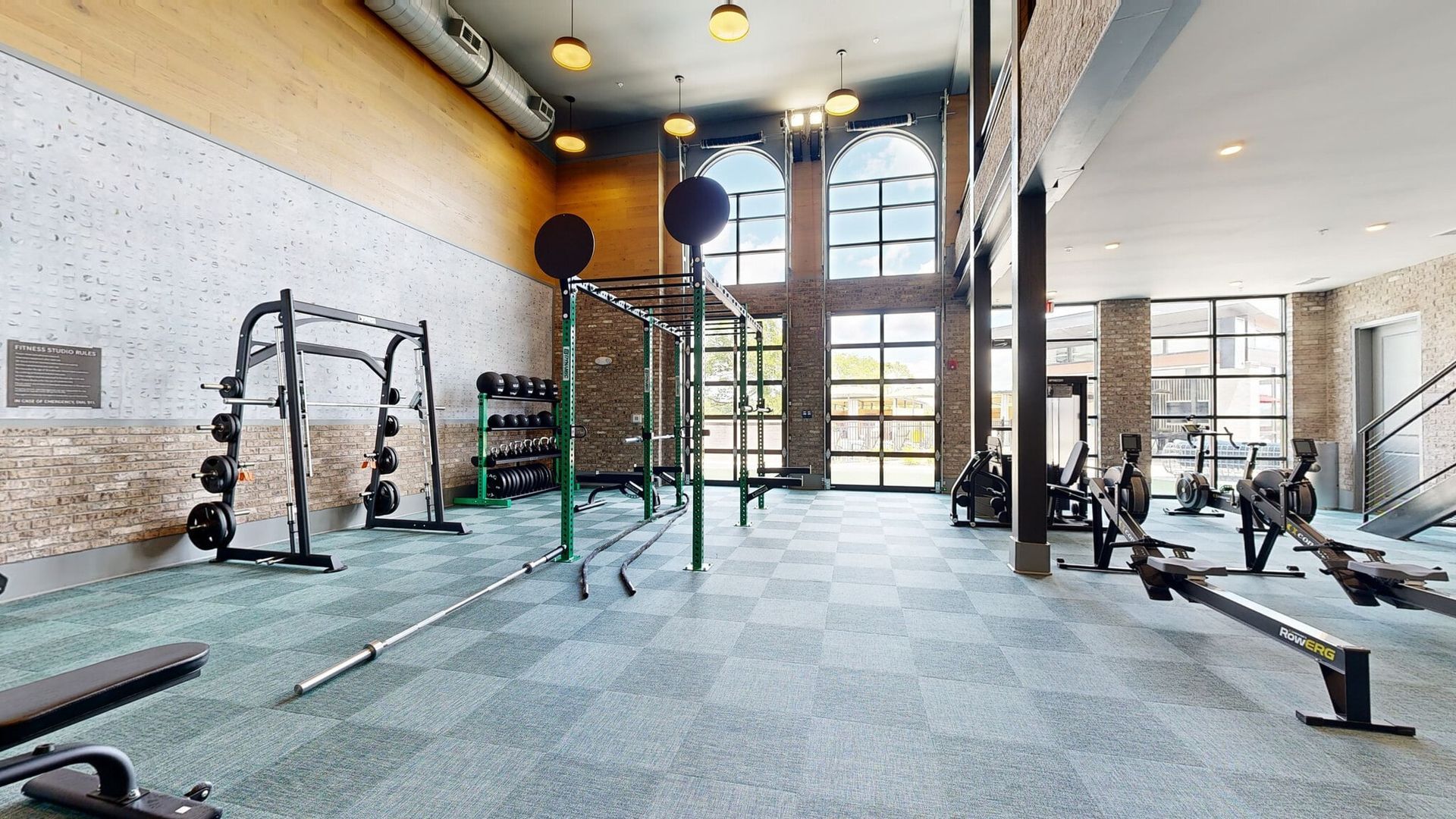 Bright gym with free weights, racks, barbells, benches, and cardio machines near tall arched windows.